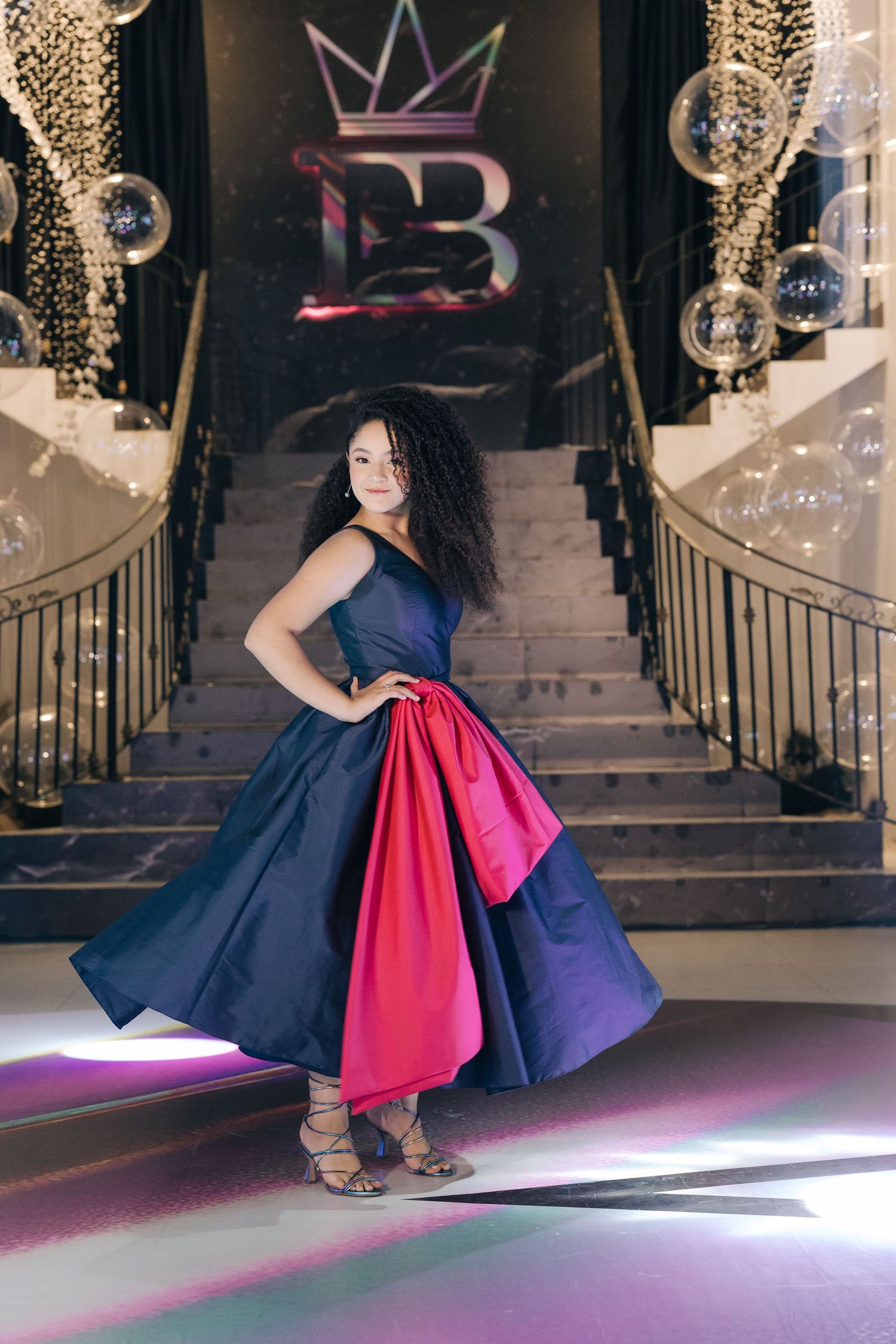 A full-length portrait on stairs showcasing a striking navy and red formal gown with a crown logo background.