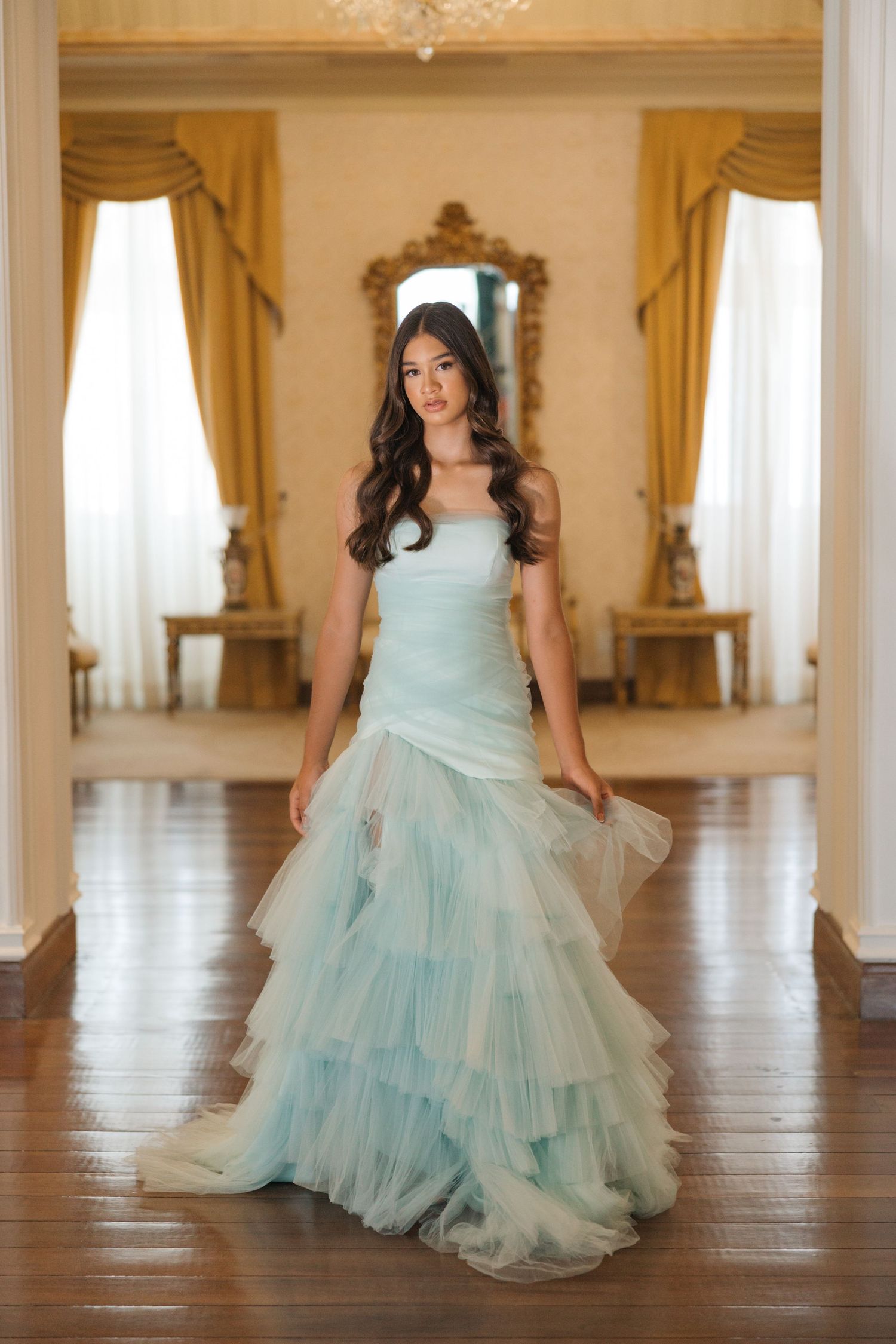 A dreamy light blue tulle gown with tiered ruffles is displayed in an elegant room with golden accents.