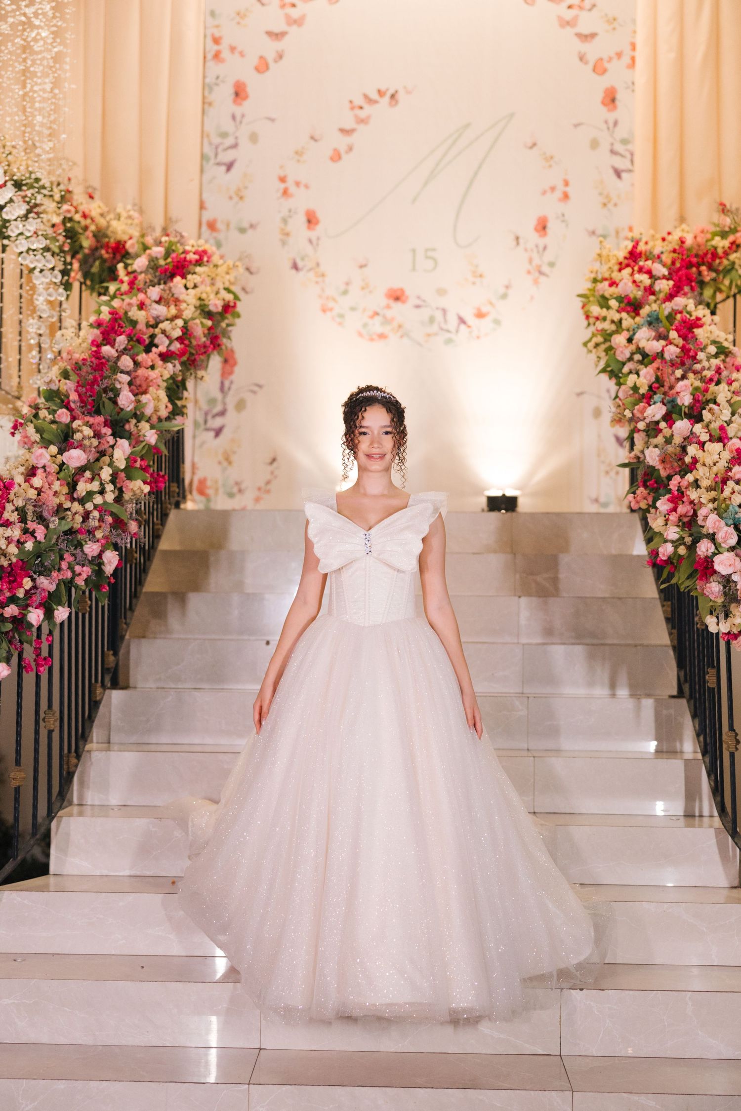 A bride in a white ballgown poses on grand staircase decorated with pink and coral floral arrangements.