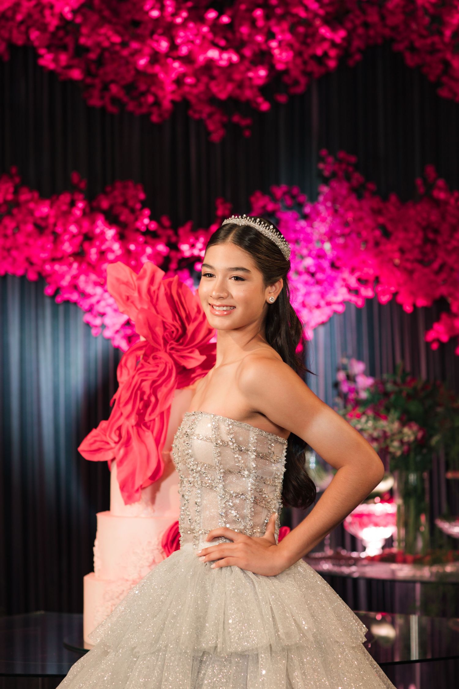 A sparkly strapless ballgown is showcased against a backdrop of bright pink floral decorations.