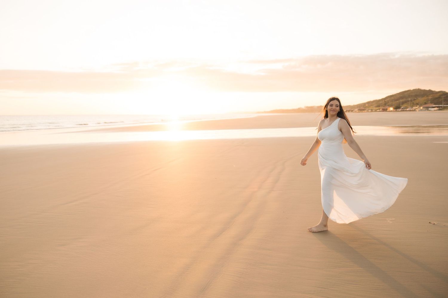 A figure in a white dress twirls on a beach at golden hour as the sun sets over the horizon.