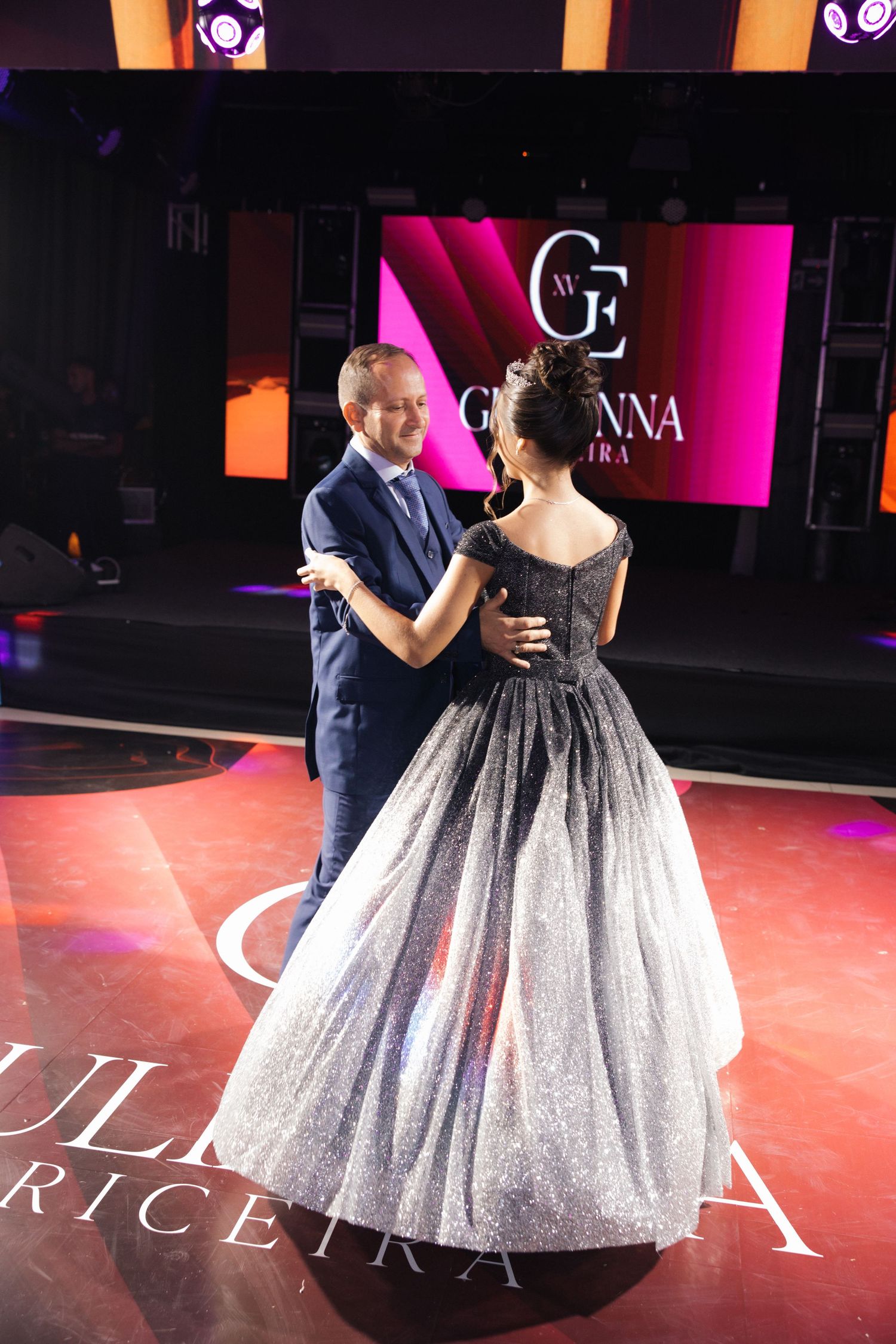 A couple dancing at an elegant event with a sparkling ombre ball gown against a pink-lit backdrop with illuminated signage.