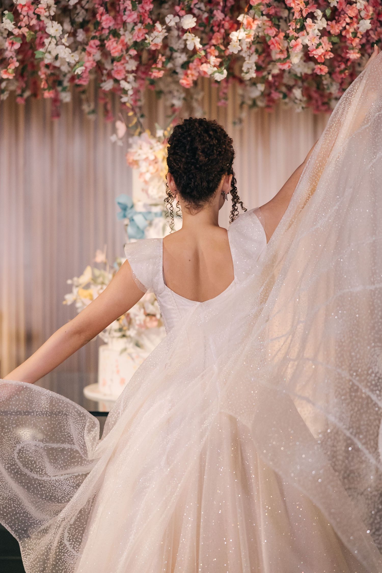 Back view of a wedding dress showing intricate details and sparkly tulle against floral backdrop.