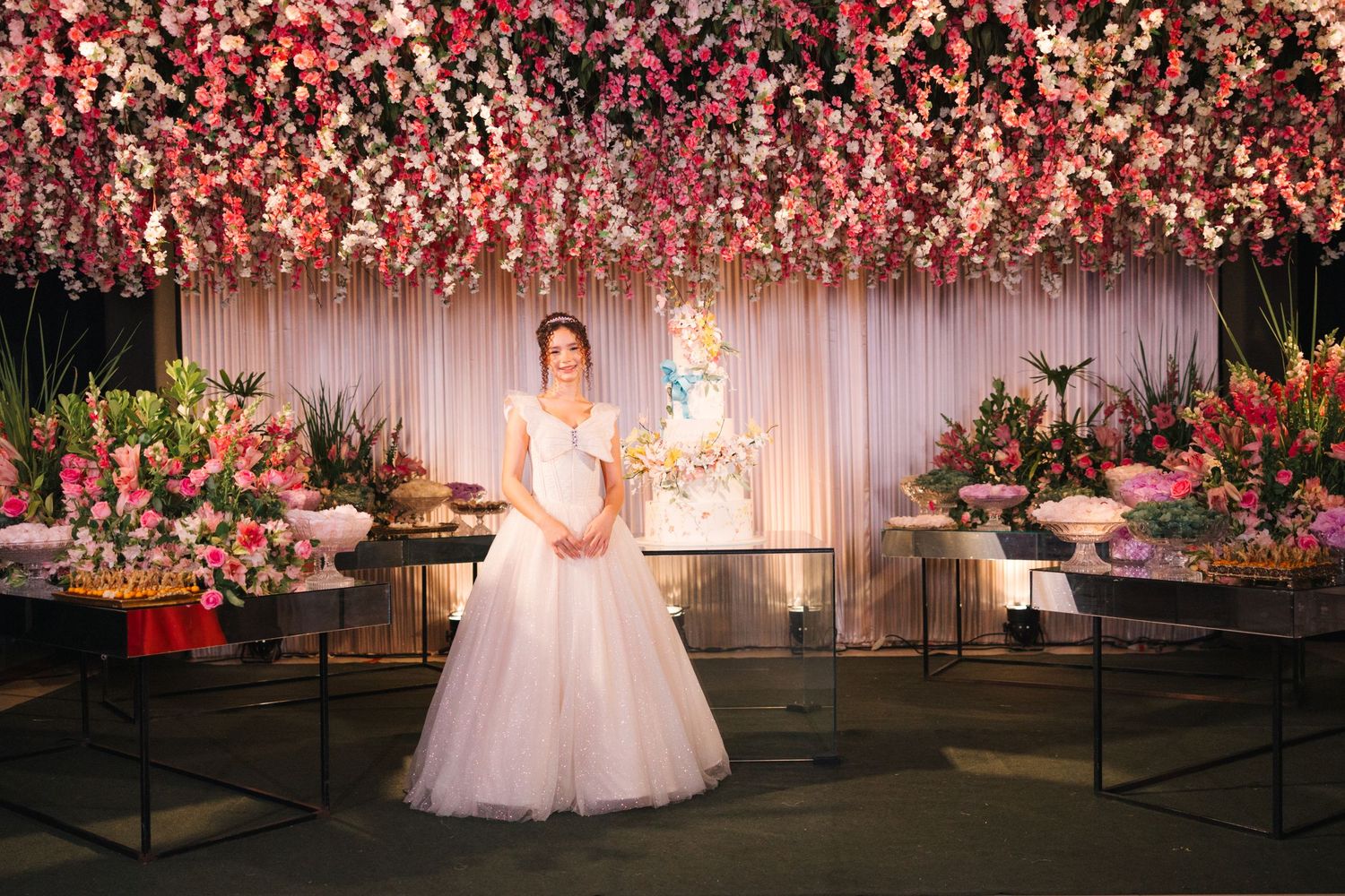 An elegant wedding reception setup with pink floral ceiling installations, dark display tables and romantic lighting.