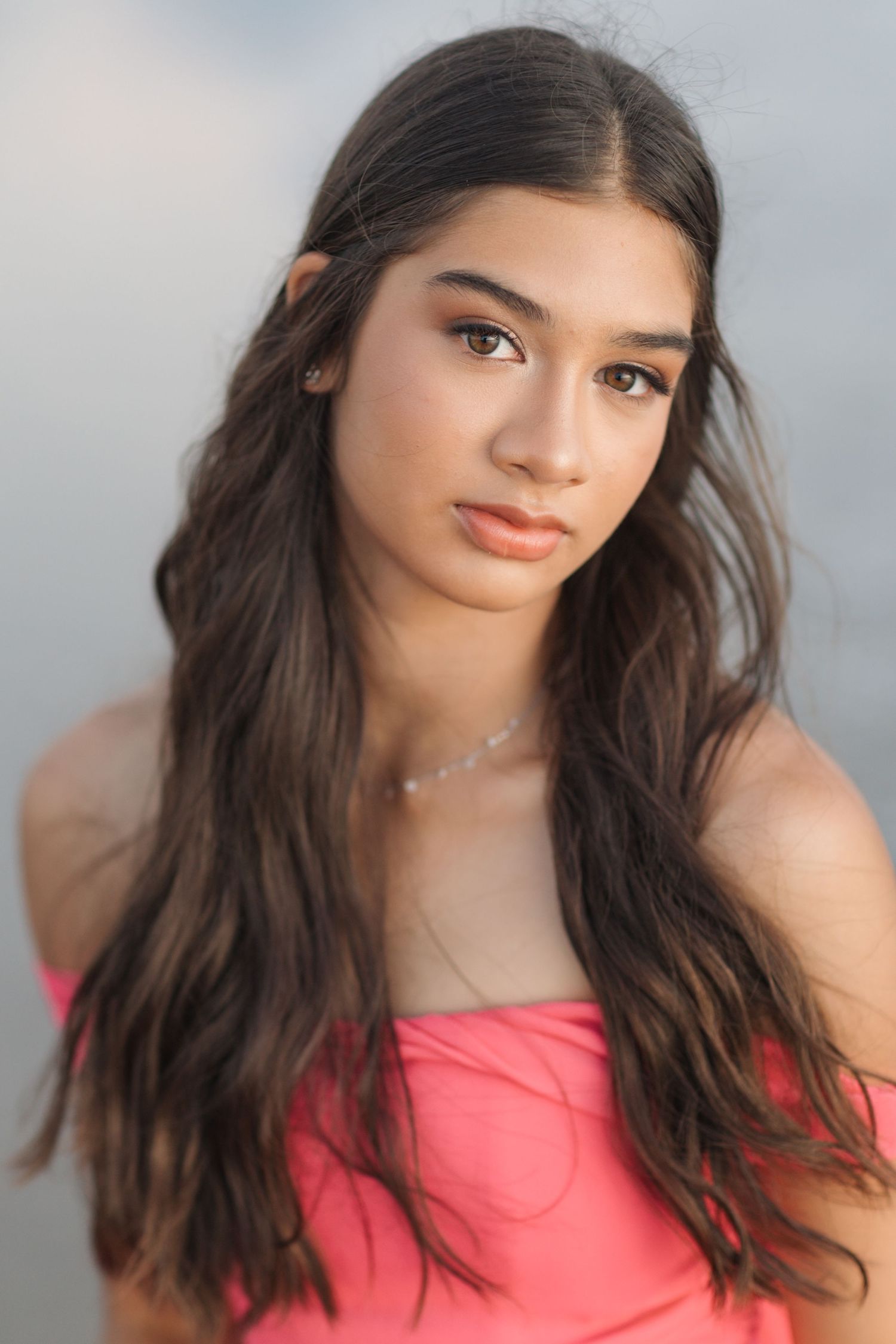 Portrait of a young woman in a coral top with long dark wavy hair against a soft blurred background.