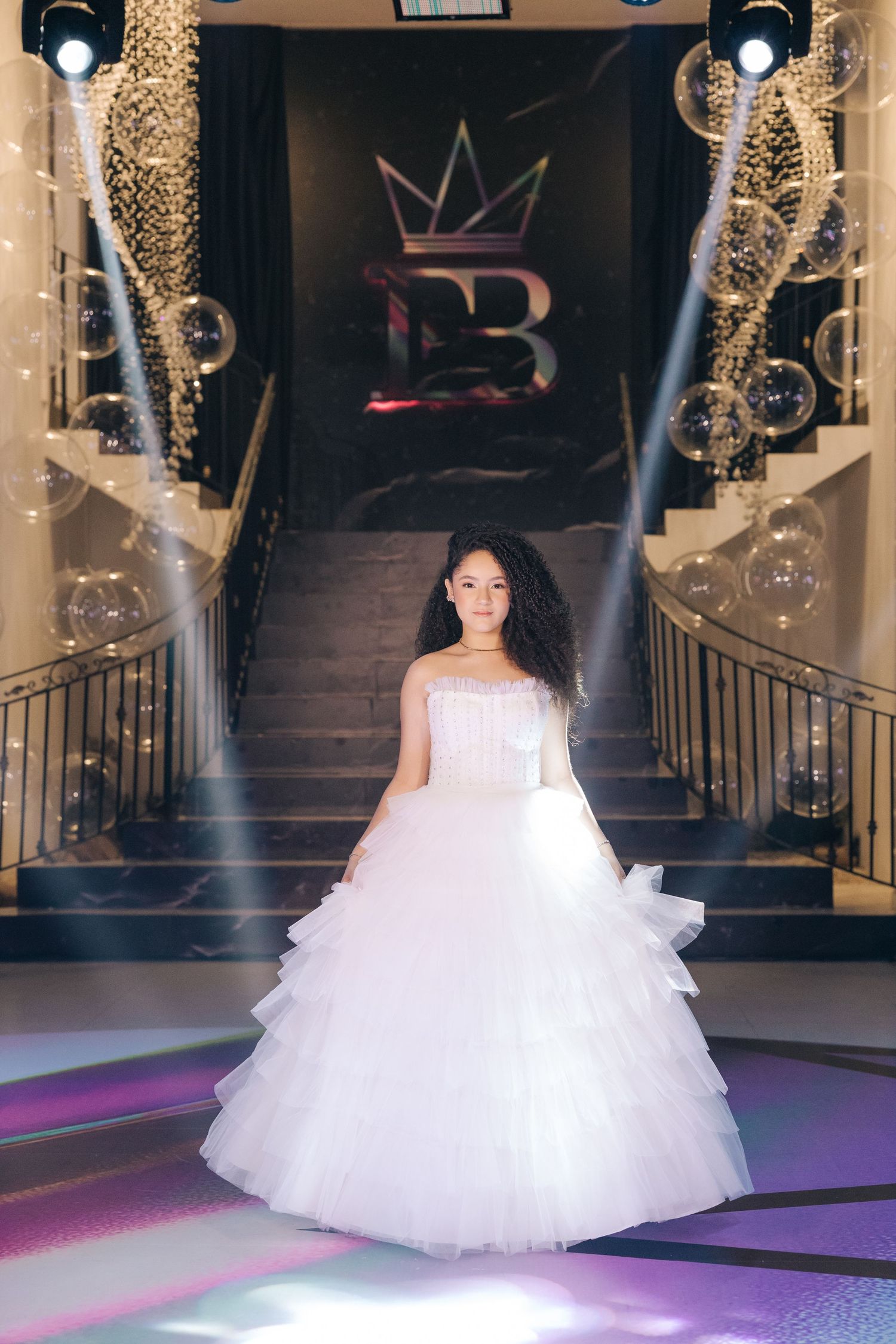 A formal white ballgown on display at an elegant venue with dramatic lighting and a crown logo.