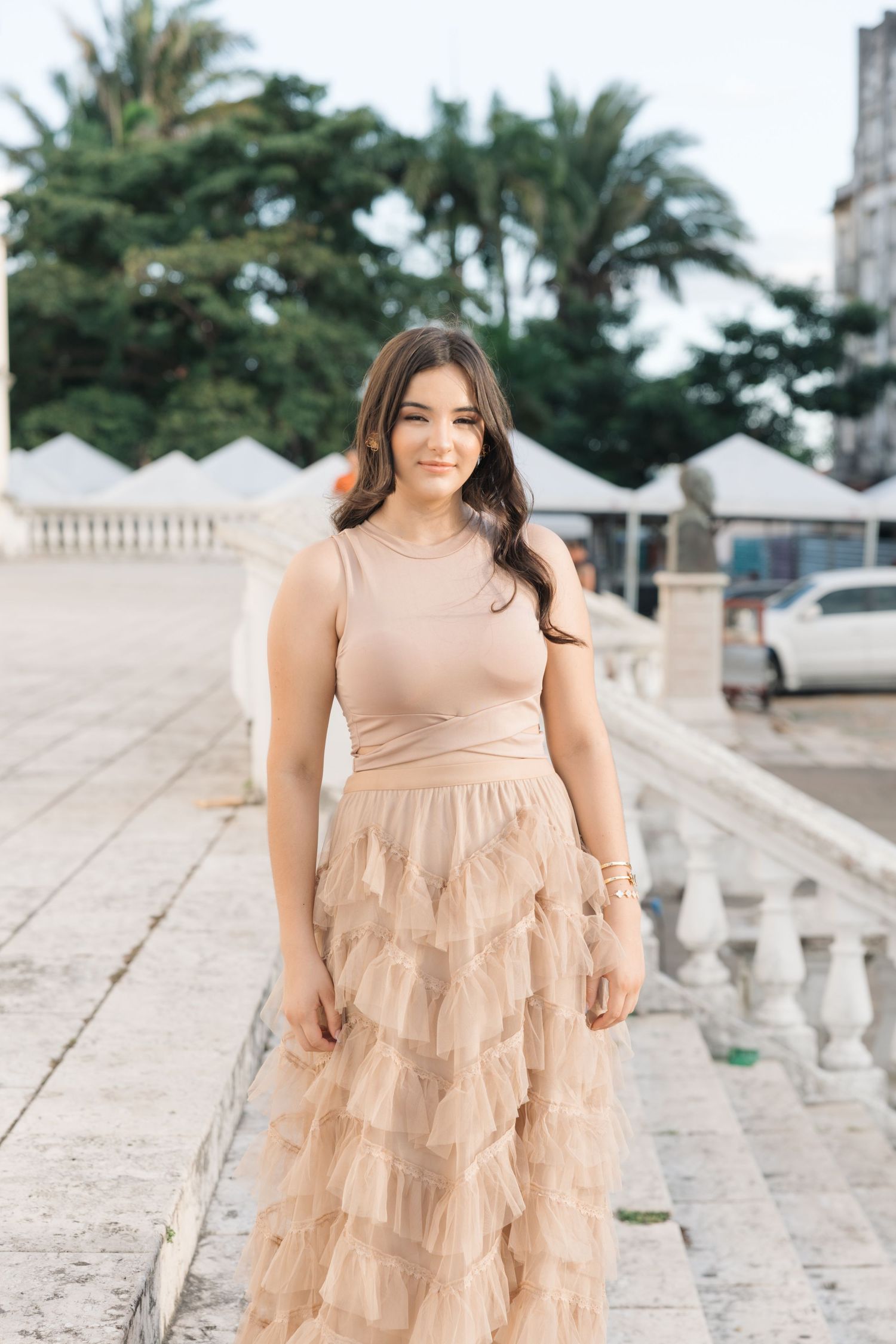 Woman in beige sleeveless dress with tiered ruffled skirt poses on stone pathway with palm trees in background.
