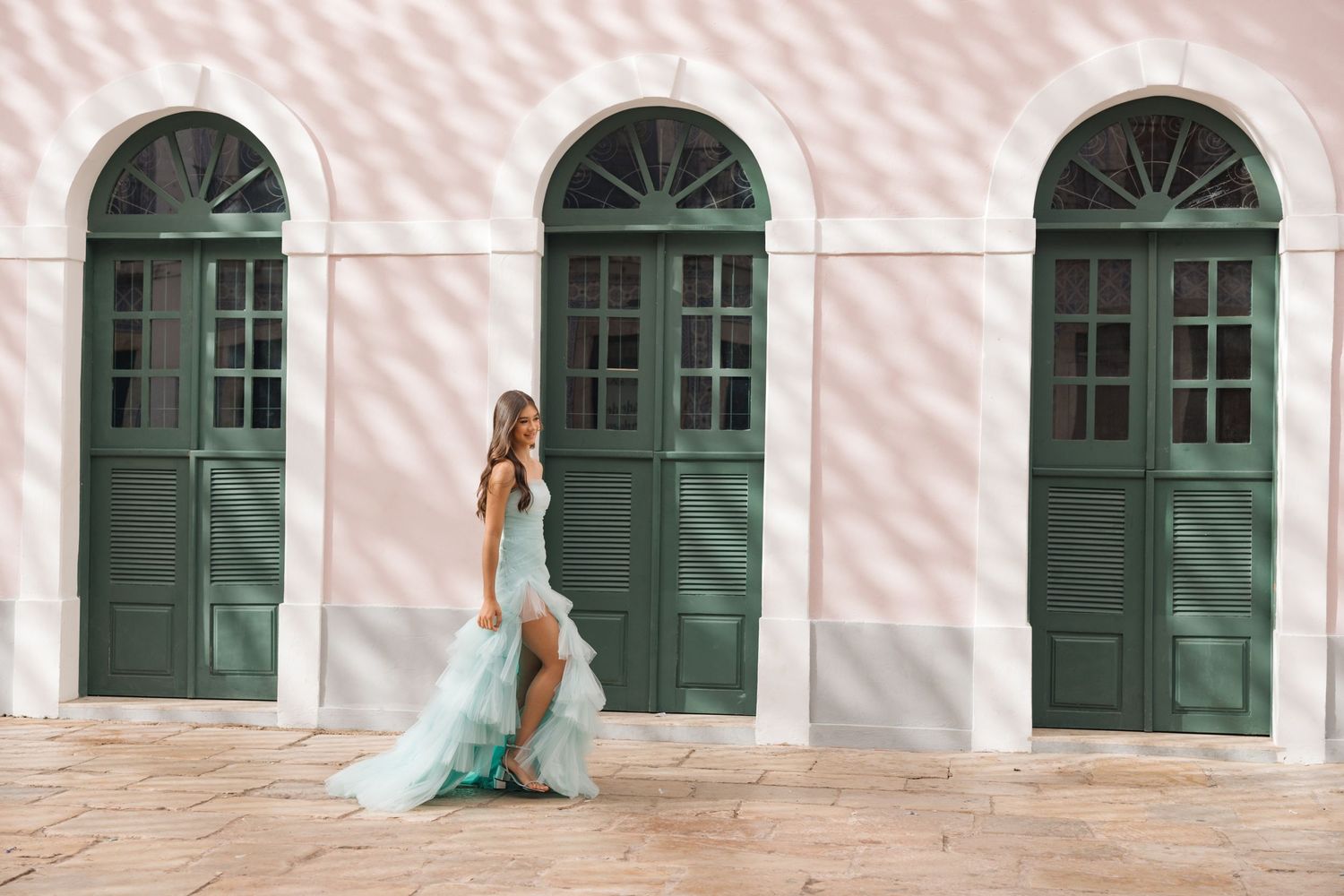 A person in a flowing turquoise dress poses against a pink wall with three green arched doorways.