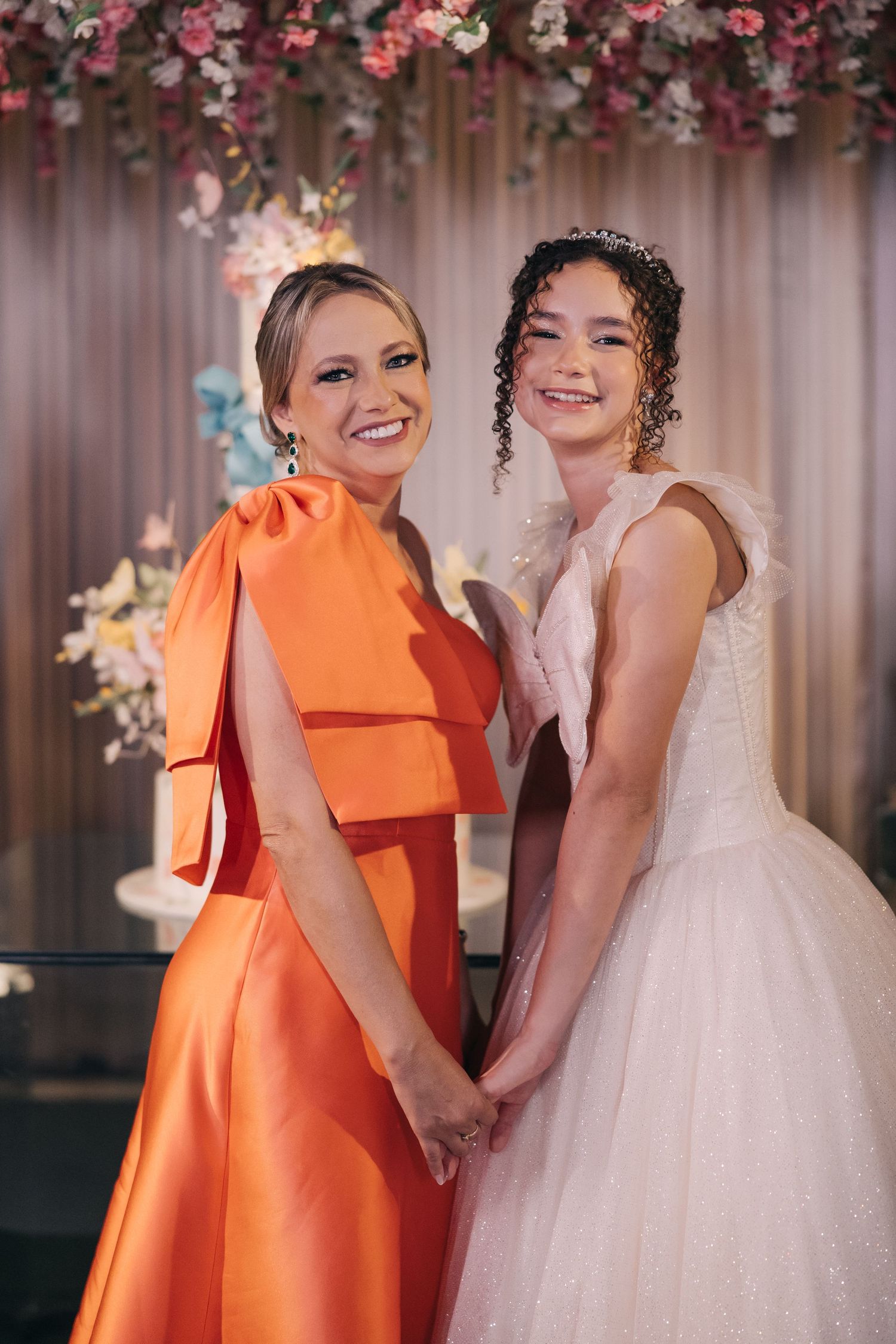 Two people posing together at a formal event, one in orange formal dress and one in white wedding gown.