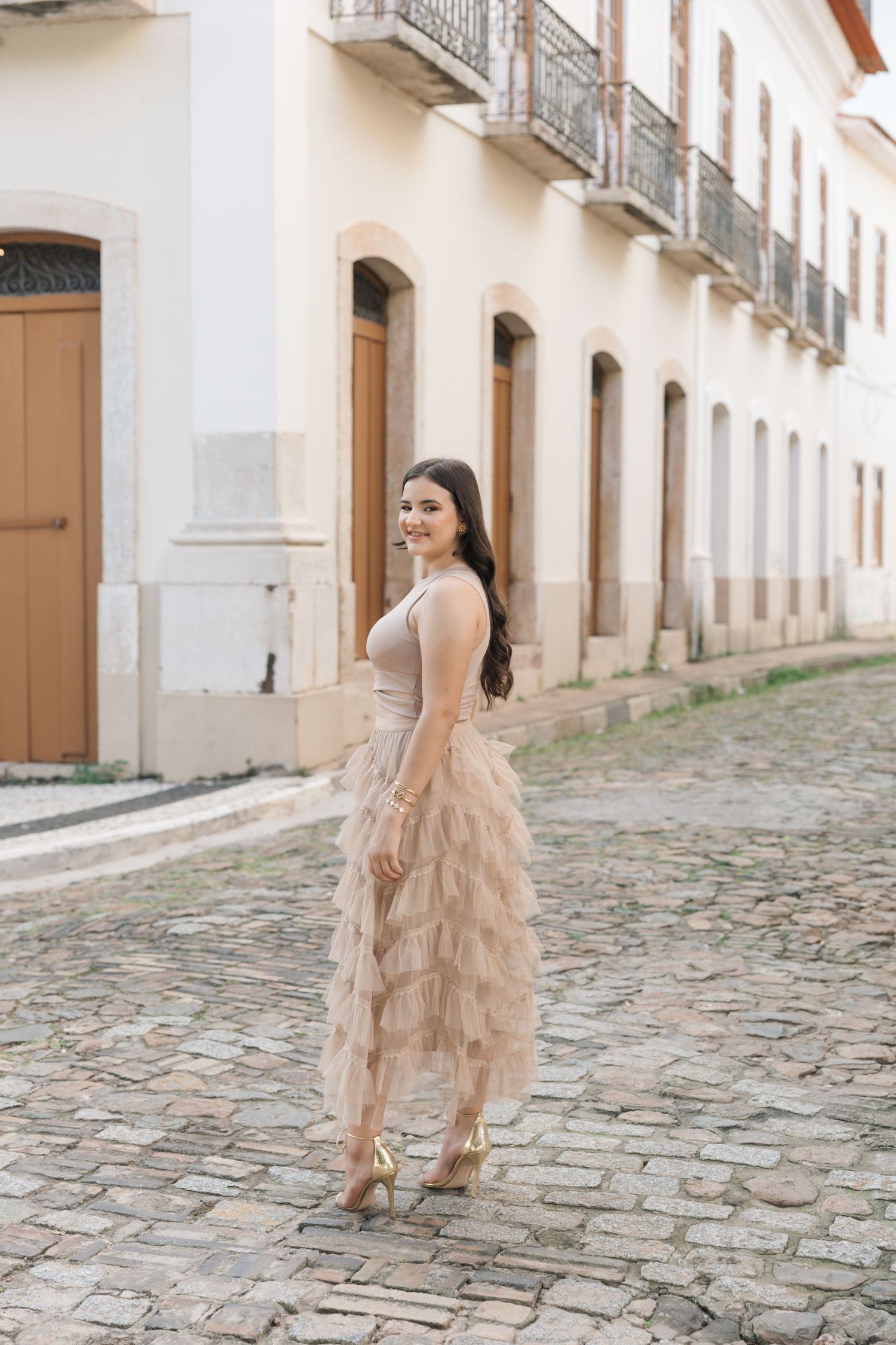 Woman in delicate tulle dress poses on cobblestone street with white colonial buildings in historic town setting.
