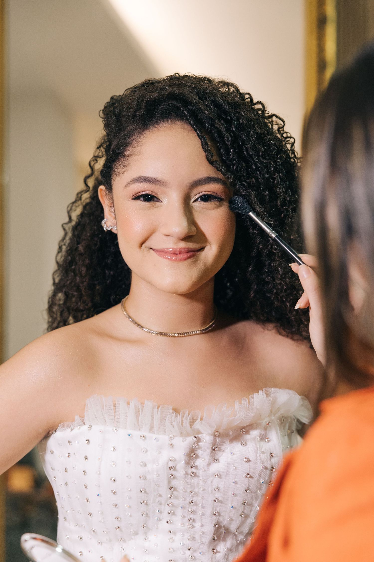 A young woman in a white gown getting her makeup done while wearing a delicate necklace.