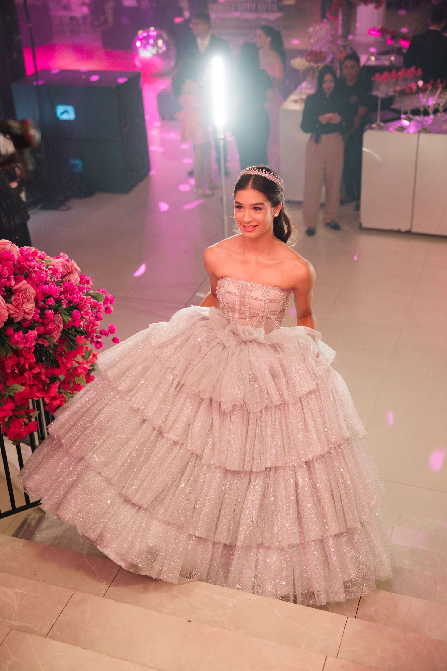 A sparkly tiered ballgown in a pink-themed party space with bright floral arrangements and ambient lighting.