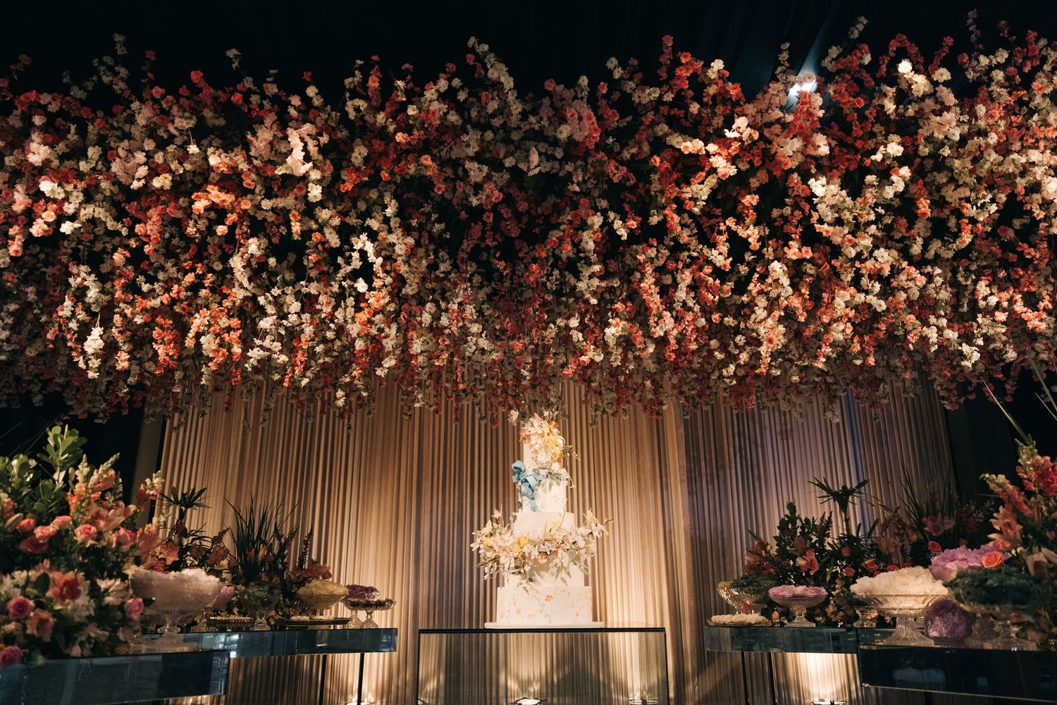 Stunning floral ceiling installation with illuminated wedding cake display beneath.