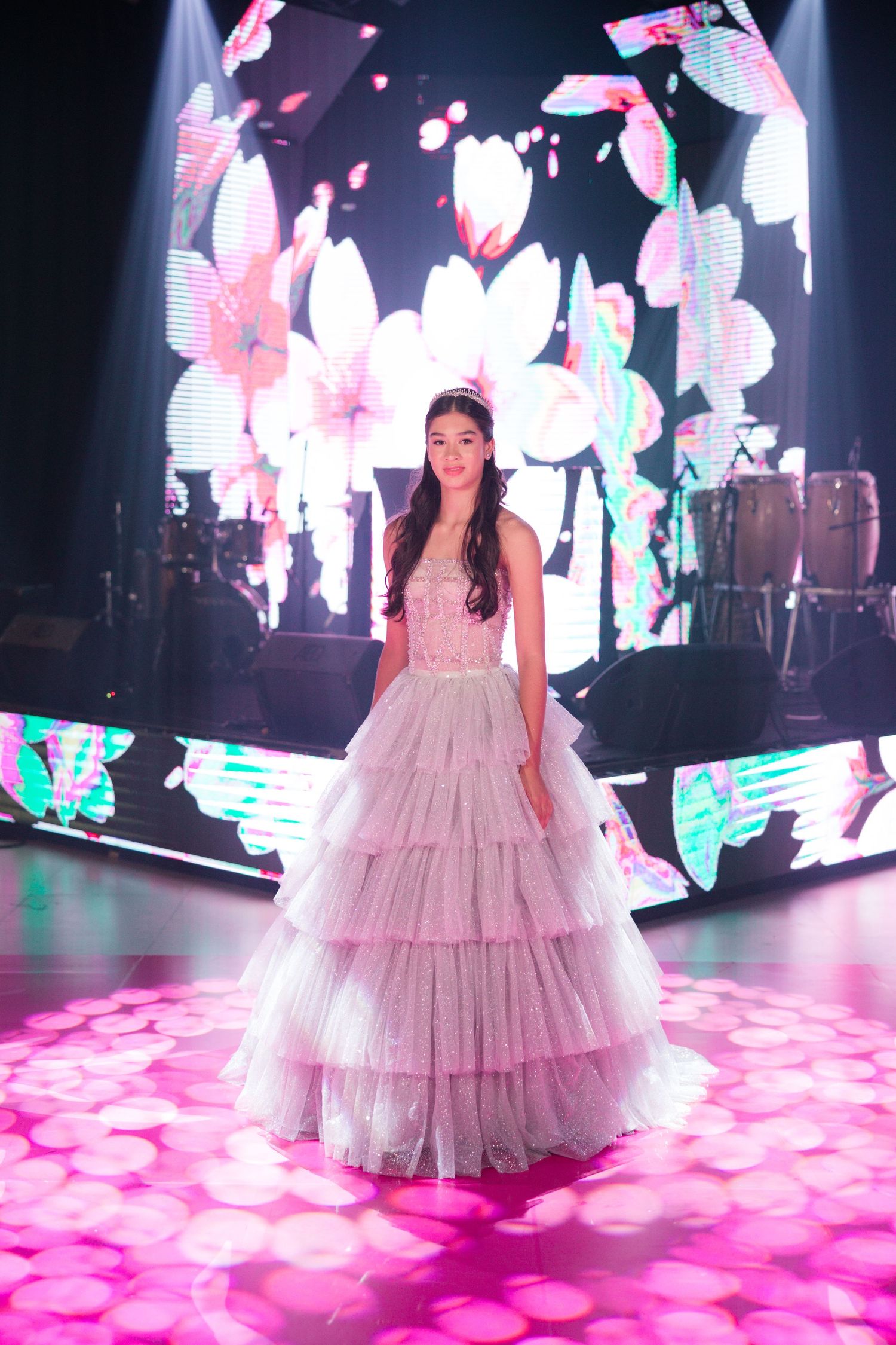 A layered tulle gown sparkles on a dance floor beneath pink floral projections and dramatic stage lighting.