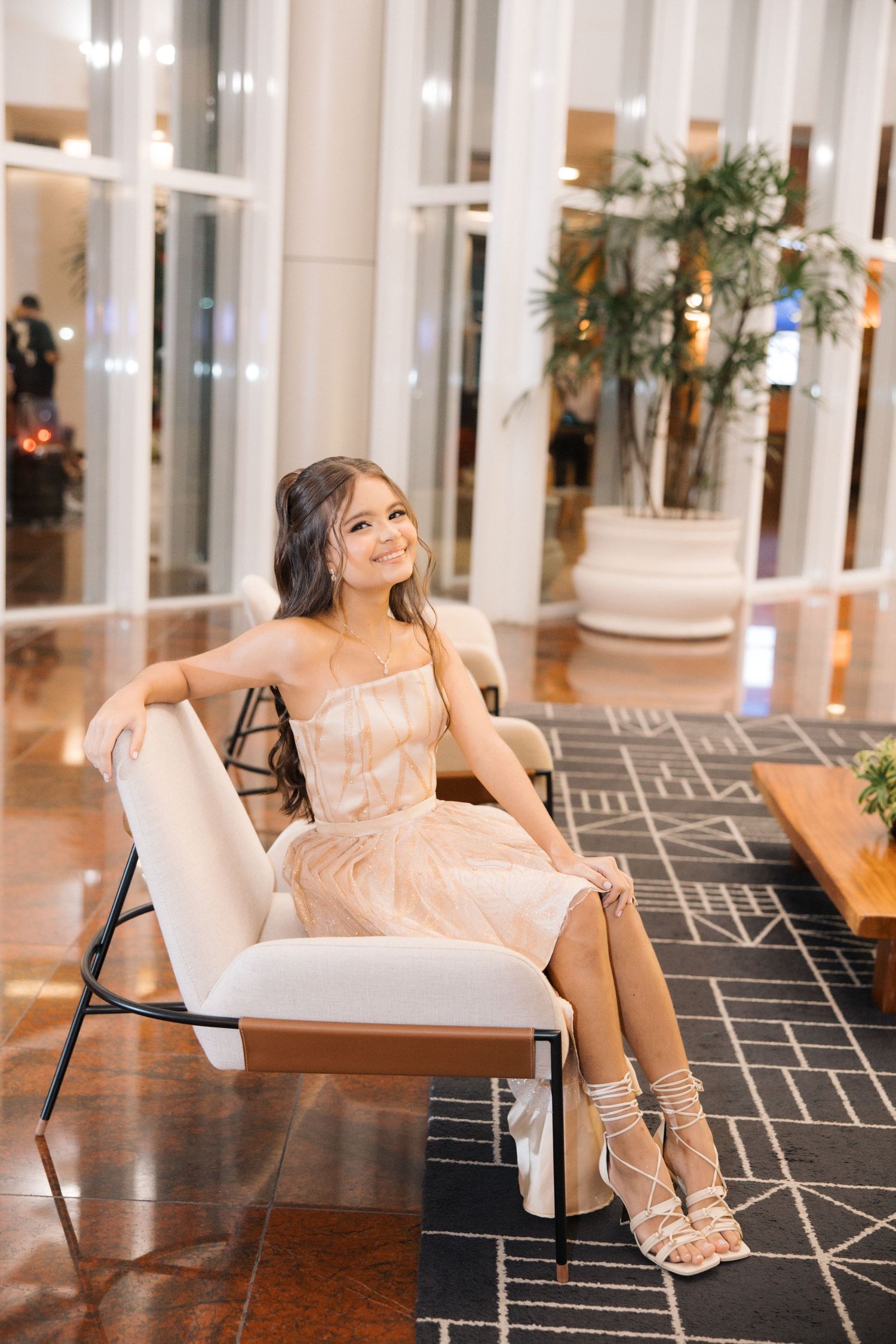 Woman in blush cocktail dress sits elegantly on white chair in modern lobby with geometric floor pattern.