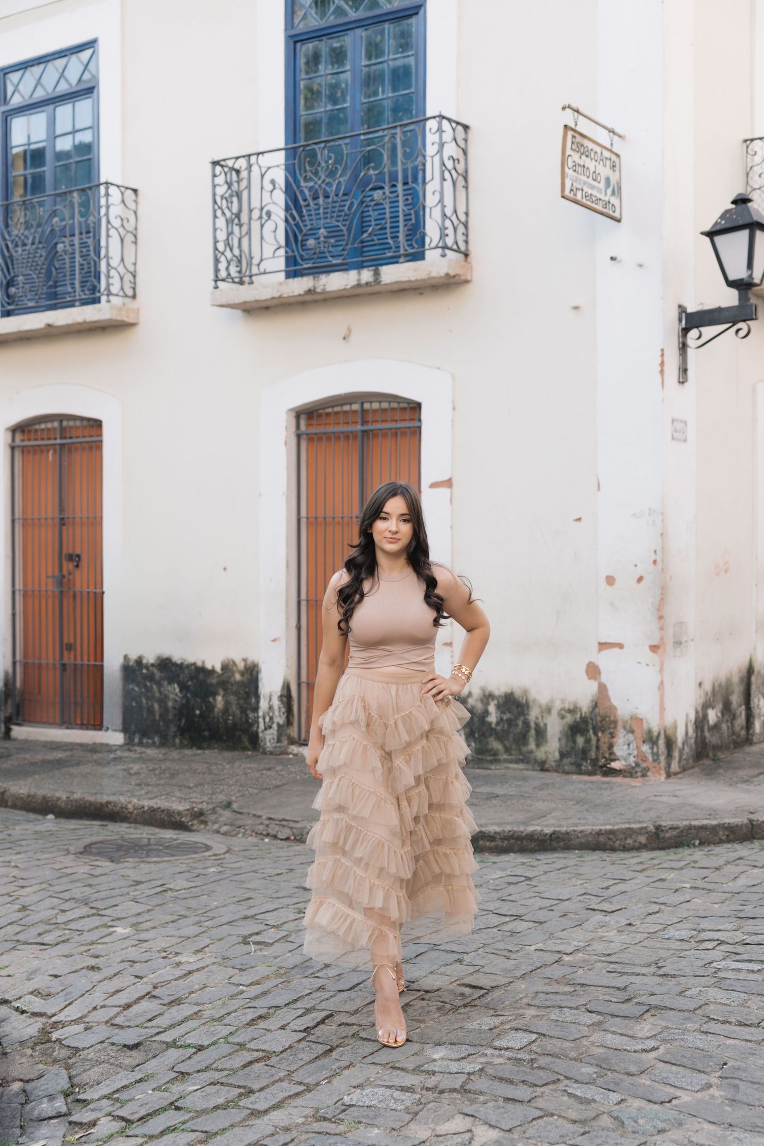 Someone in a blush-colored ruffle maxi dress poses on a historic cobblestone street with colonial architecture behind them.