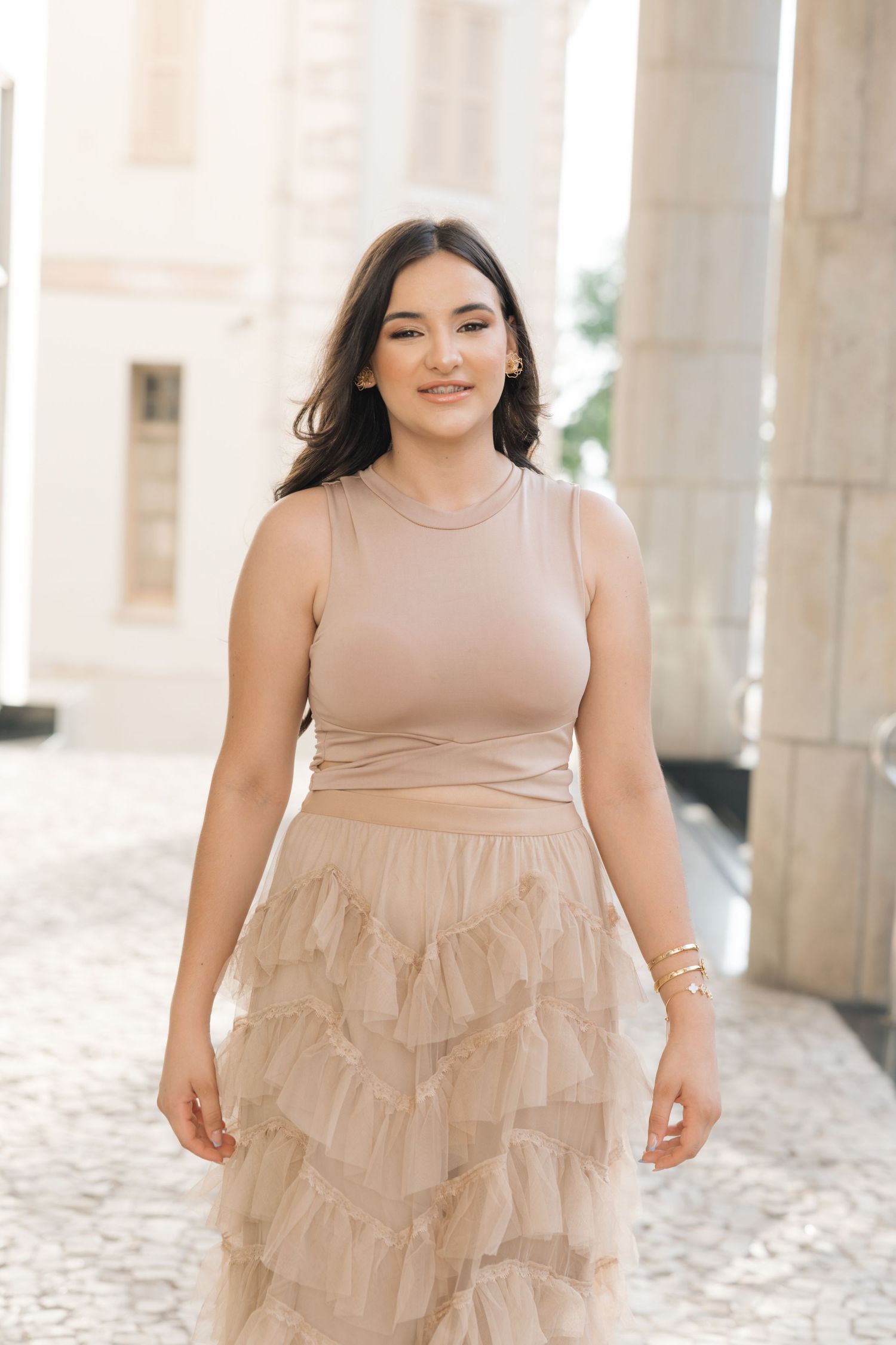 Woman in beige sleeveless top and tulle skirt poses elegantly against white pillars on a sunny day.