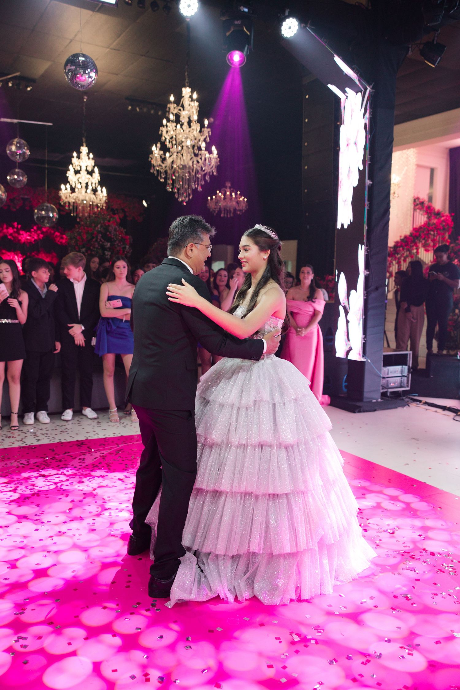A quinceanera first dance moment under crystal chandeliers with pink lighting and floral decorations.