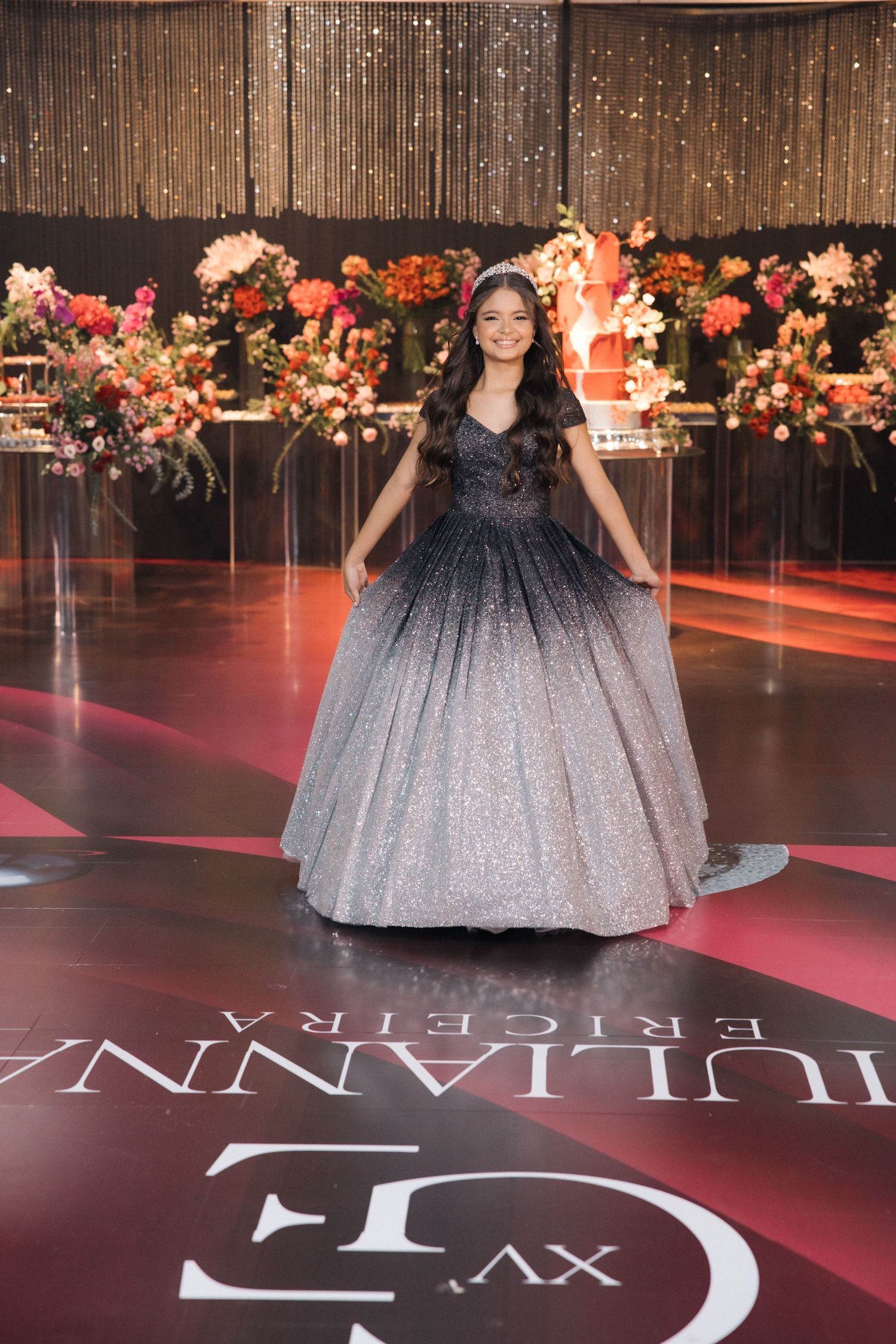 A sparkly ballgown with gradient effect from dark to light shimmers on a red carpet with floral decorations.