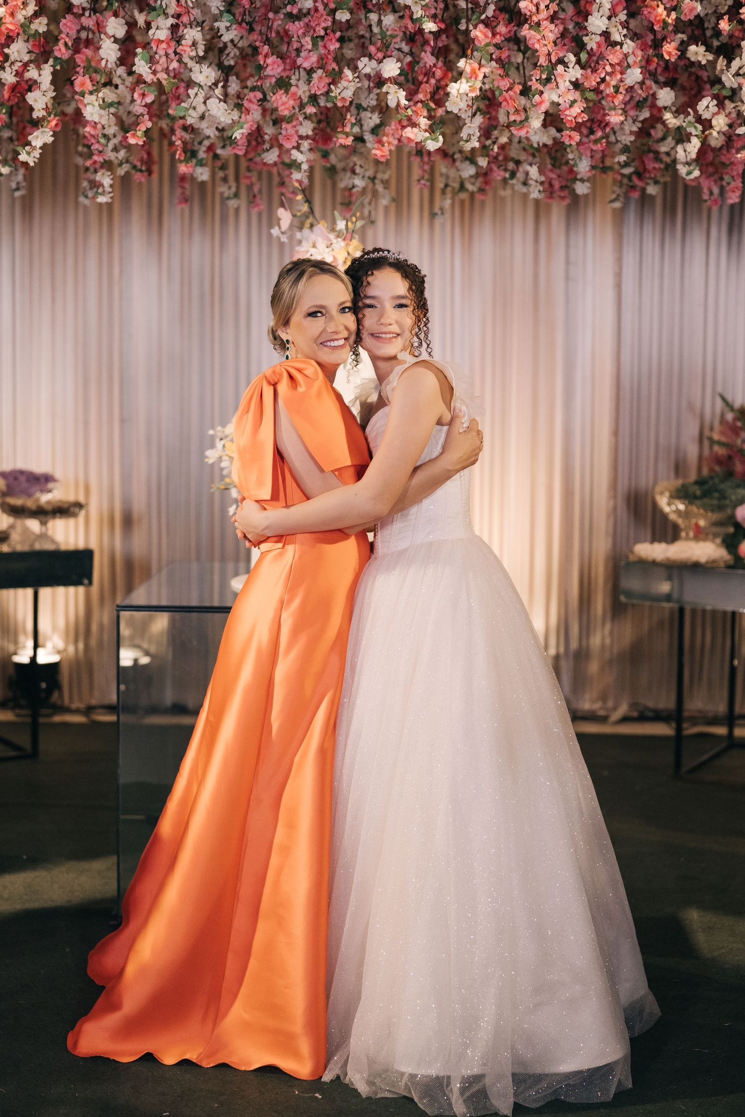 Two guests embrace at a wedding under a pink floral arch, one wearing a vibrant orange gown and one in a white ballgown.
