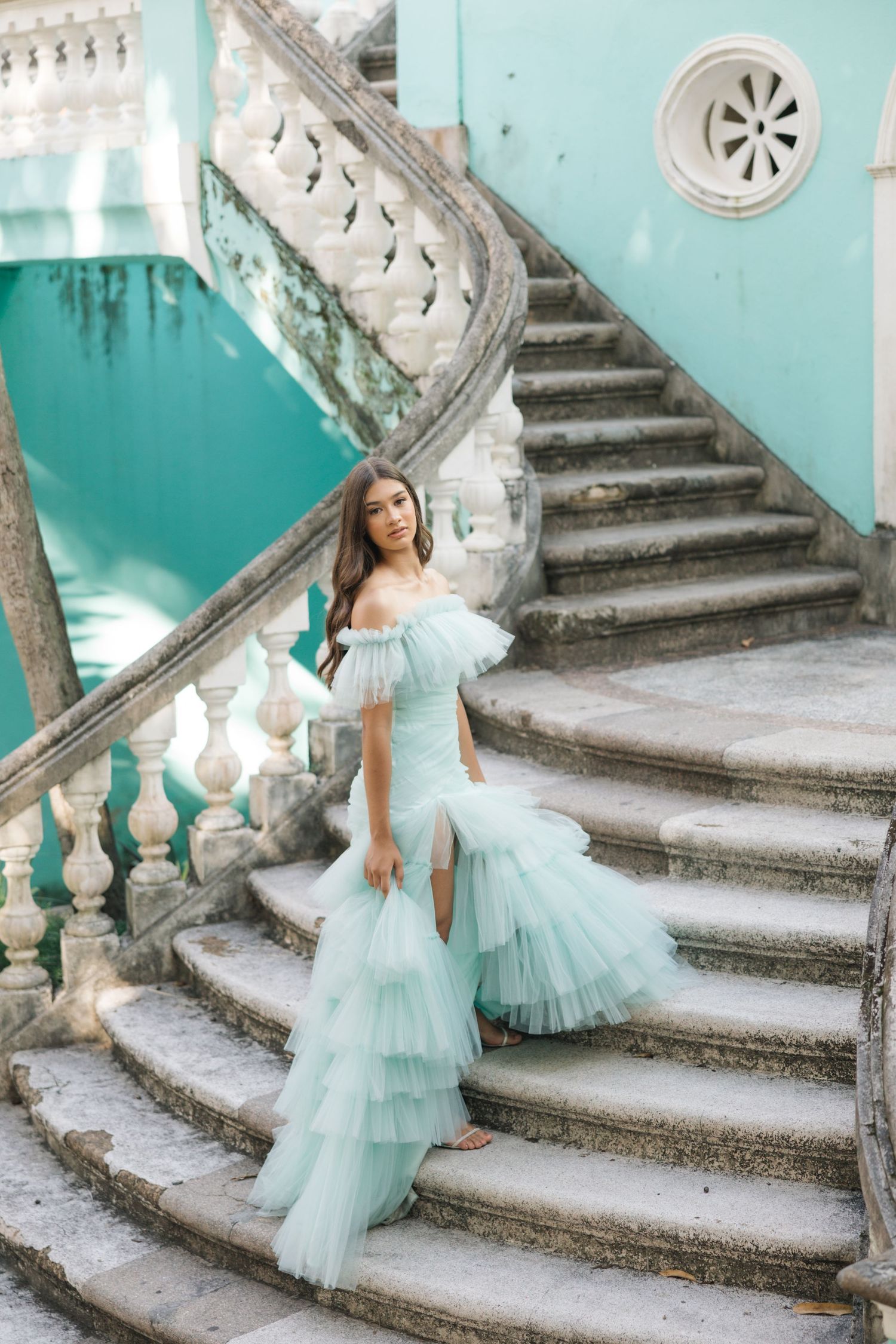 A turquoise tulle gown with tiered ruffles cascades down stone stairs against a mint-colored wall.
