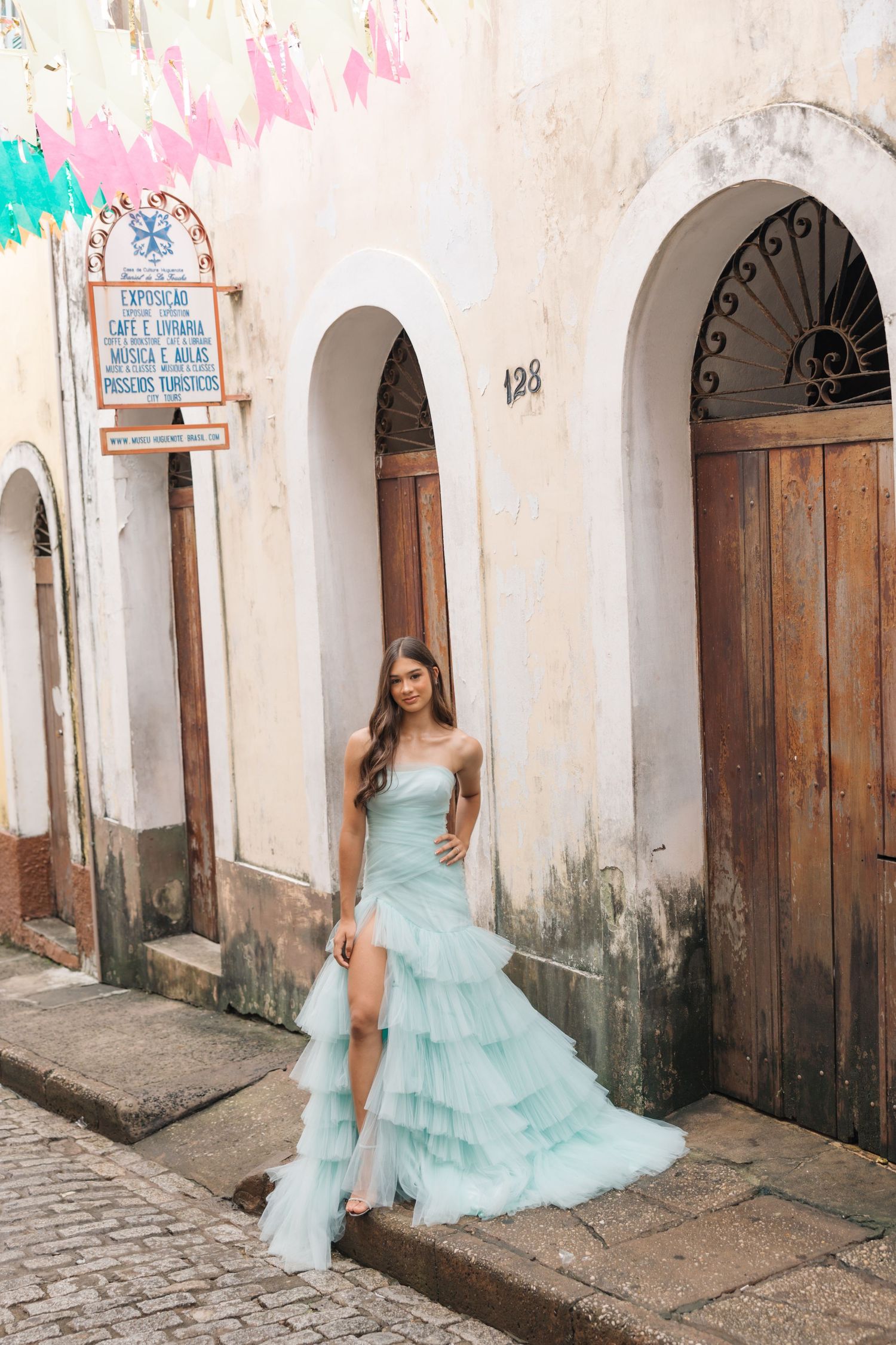 A teal ruffled gown is showcased against a historic white stucco building with wooden doors and archways.