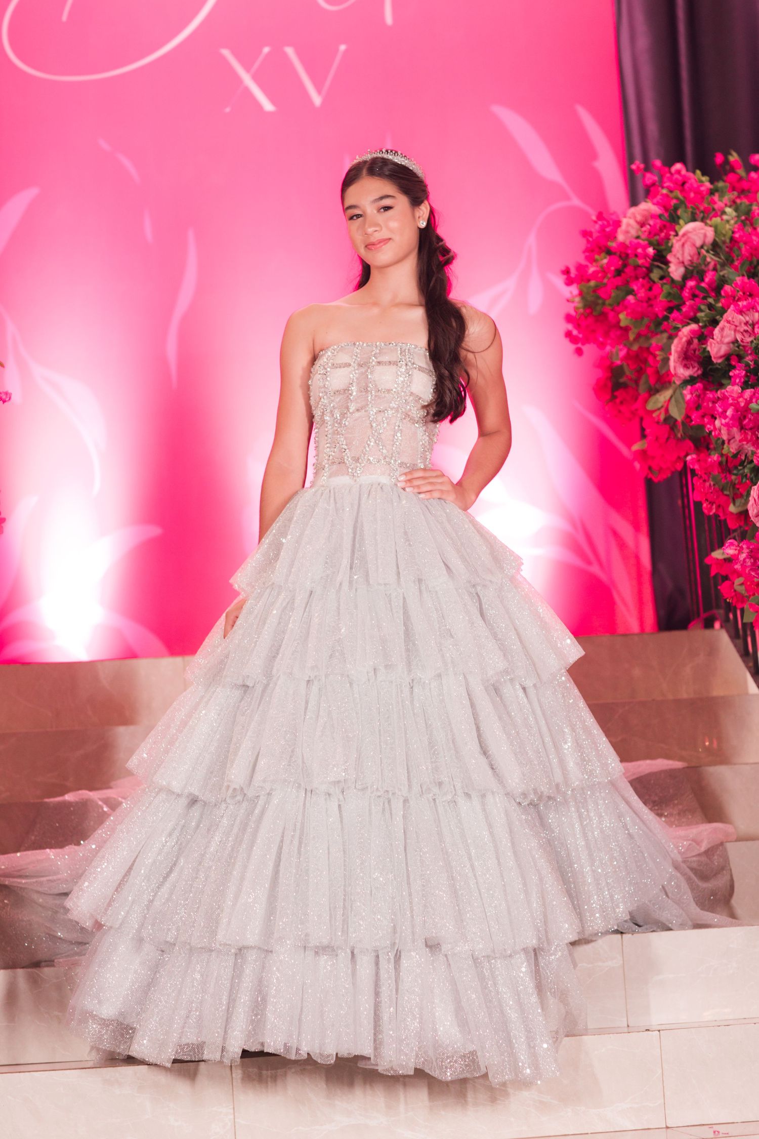 A stunning tiered white ballgown with sparkly details against a pink-lit backdrop with floral arrangements.