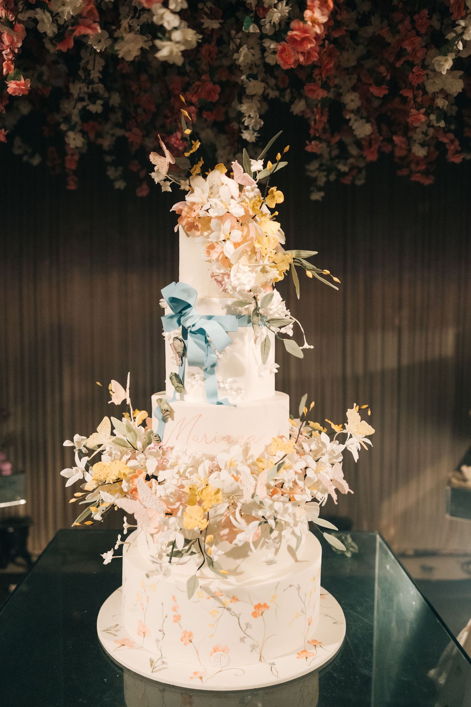 An elegant white wedding cake decorated with delicate floral arrangements and pastel accents stands against a dark backdrop.