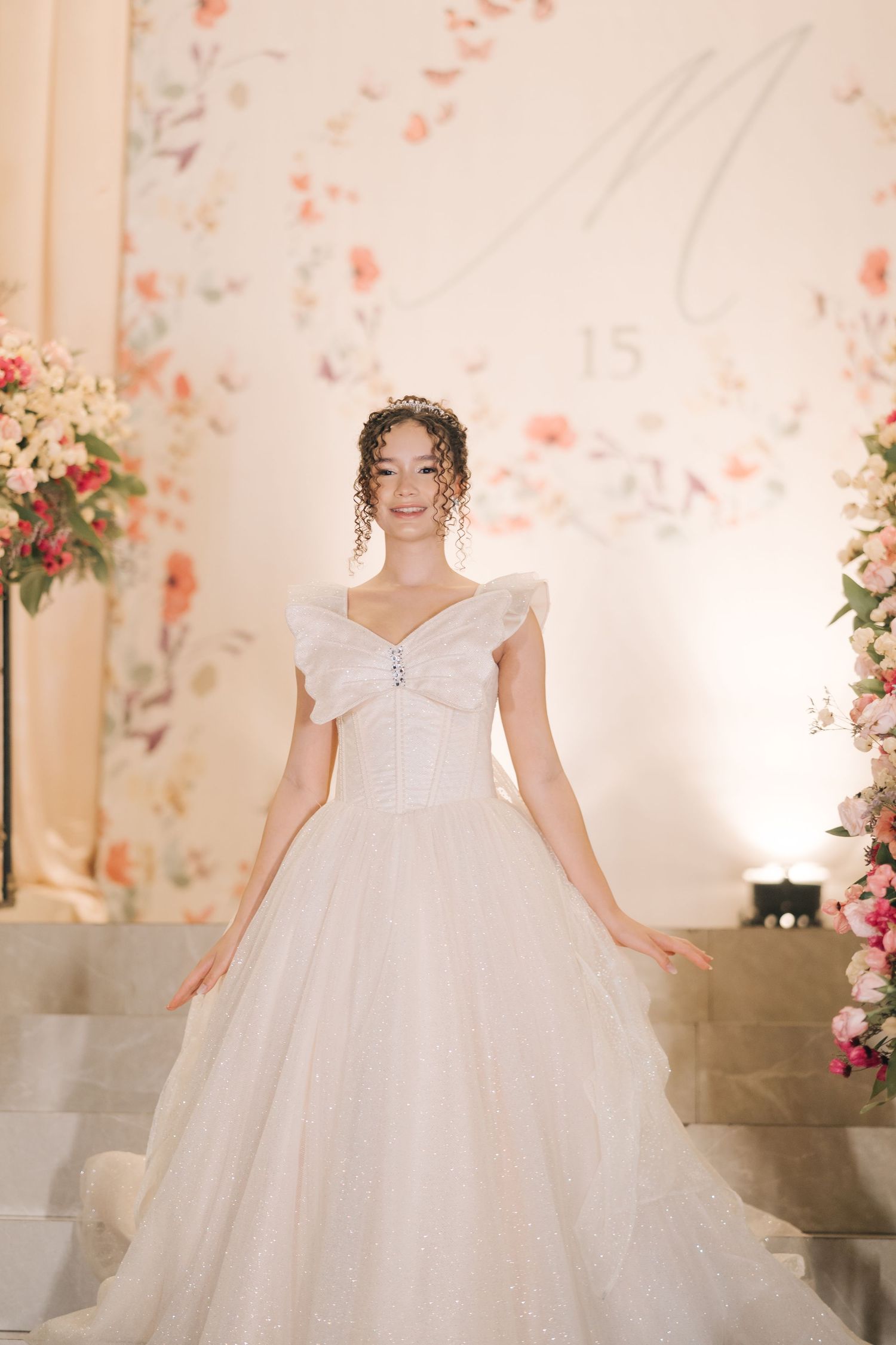 A bride poses in an elegant off-shoulder white ballgown surrounded by pink floral arrangements.