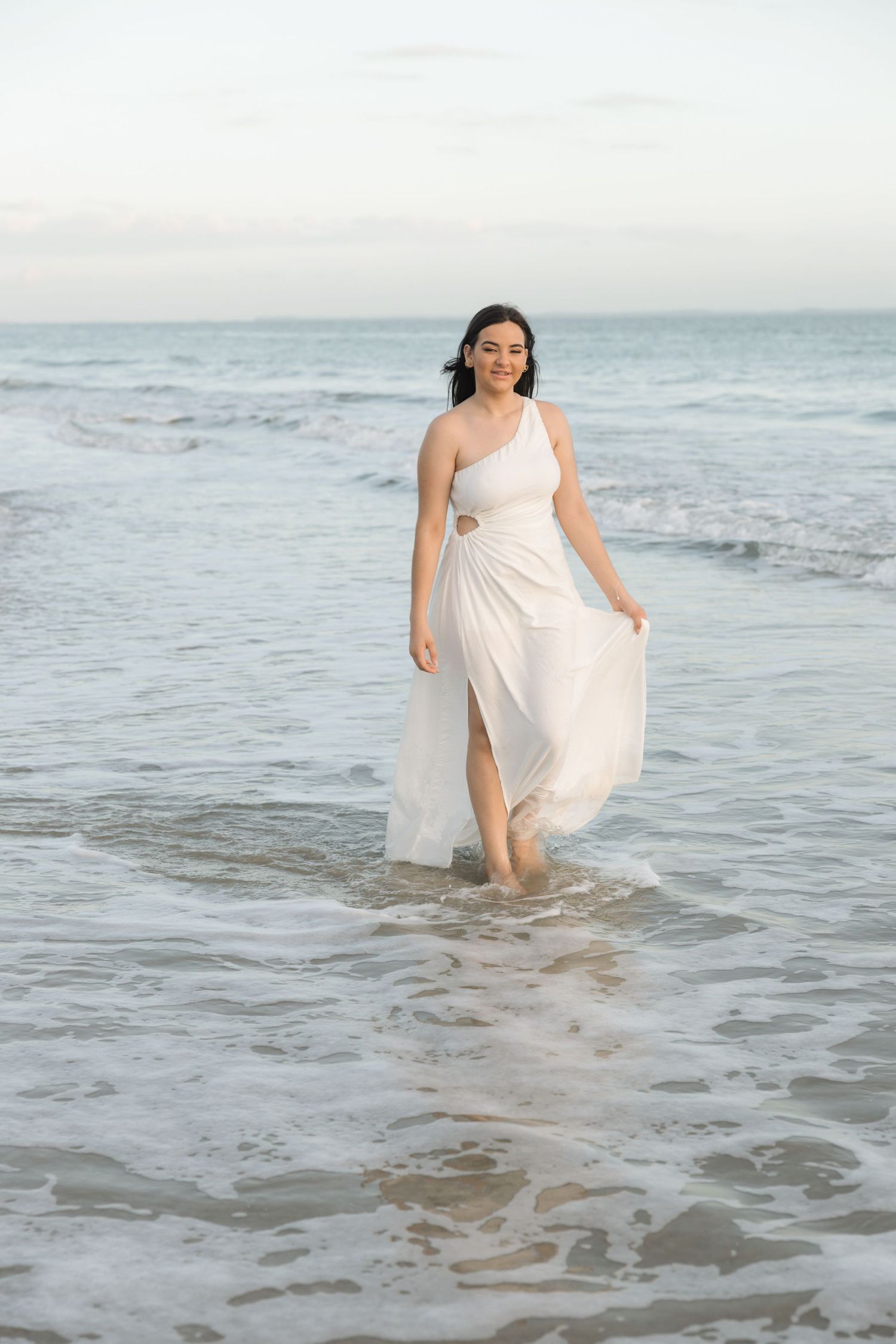 Someone wearing an elegant white one-shoulder dress walks through gentle ocean waves at the beach during sunset.