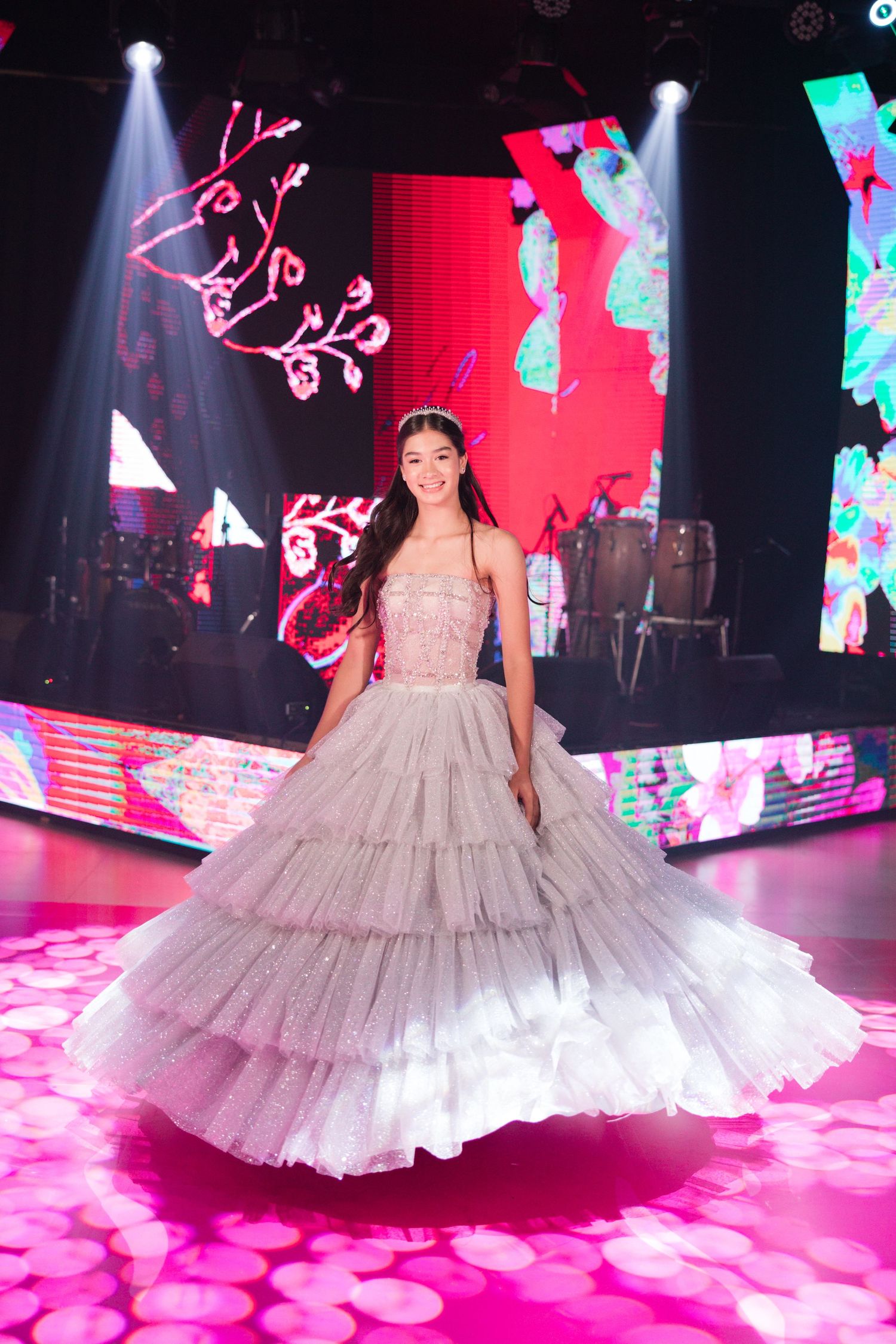 A stunning ballgown on a pink-lit dance floor with digital screens and spotlights creating a glamorous party atmosphere.