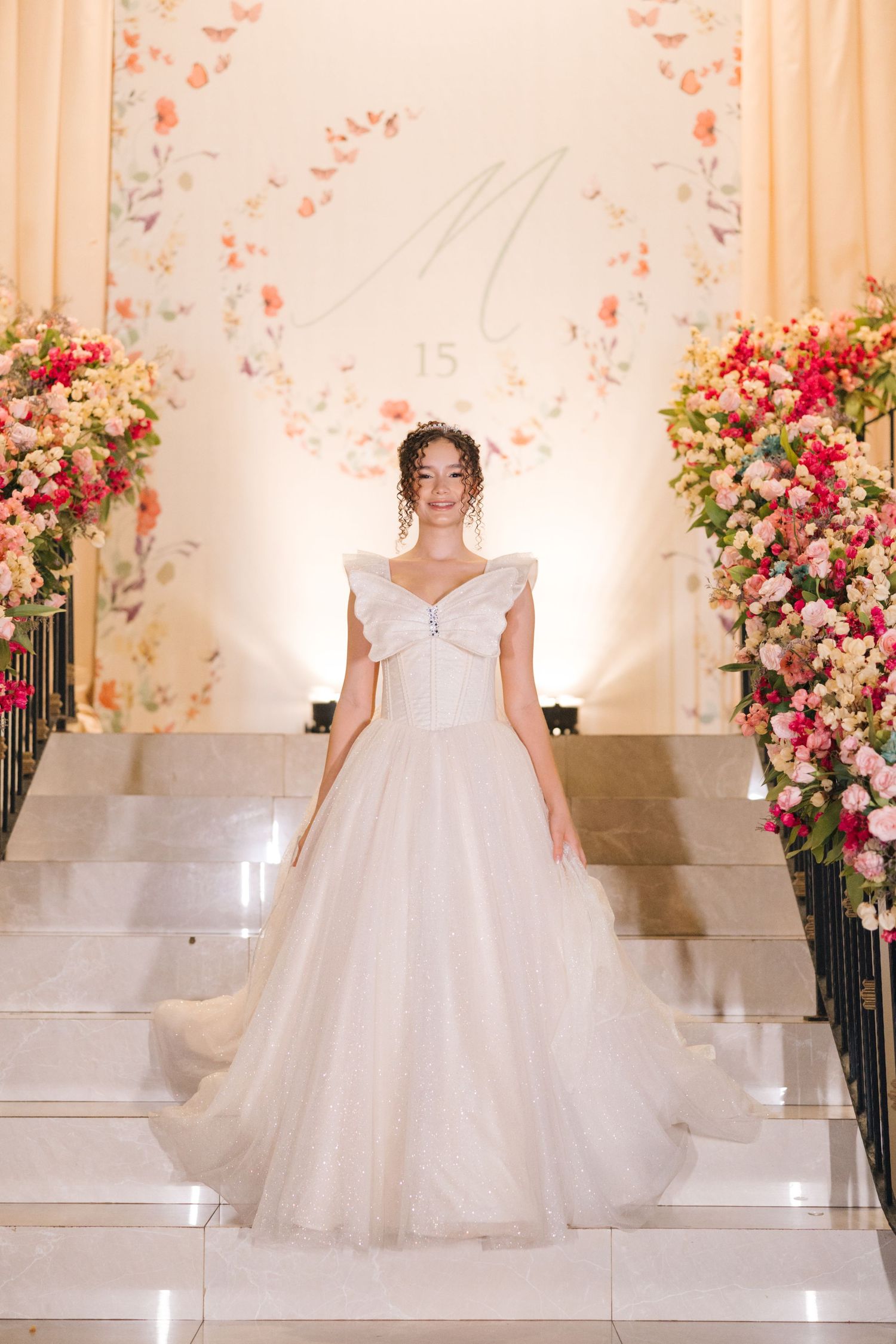 A bride in an elegant white ballgown poses on marble stairs adorned with pink and coral floral arrangements.