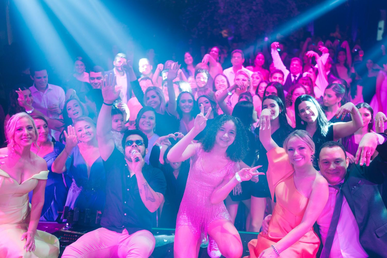 Energetic crowd dancing and celebrating at a nightclub under bright pink and blue lights.