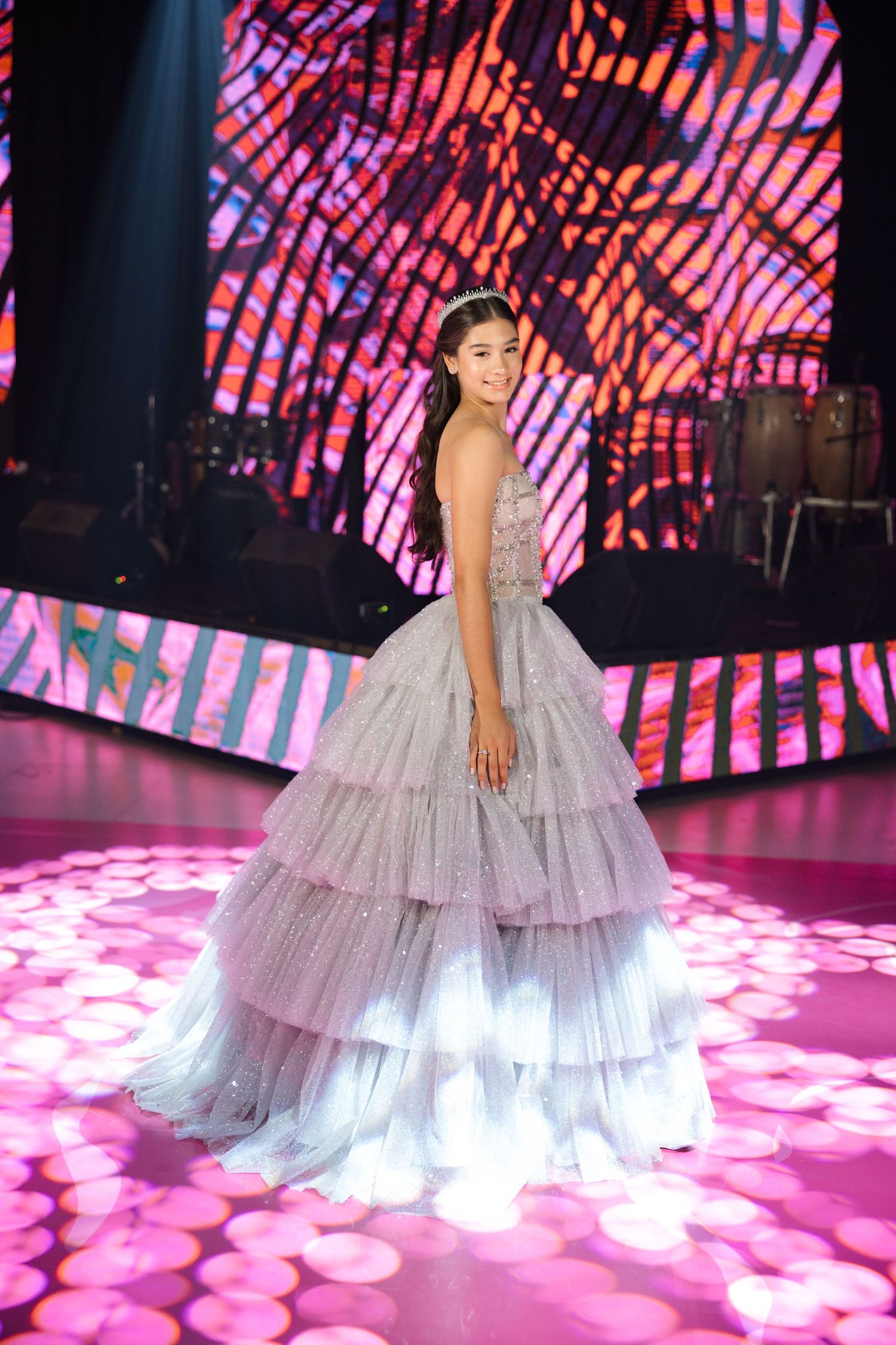 A sparkly tiered ballgown is showcased against a pink geometric backdrop with dramatic lighting effects.