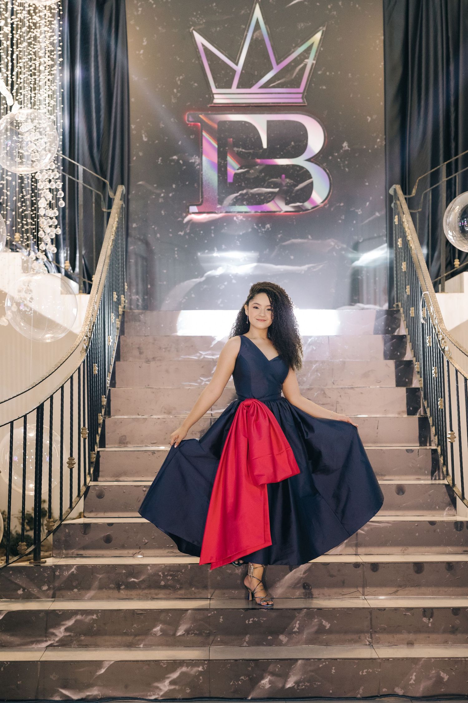A dramatic pose on elegant stairs featuring a navy and red ballgown against an illuminated crown logo backdrop.