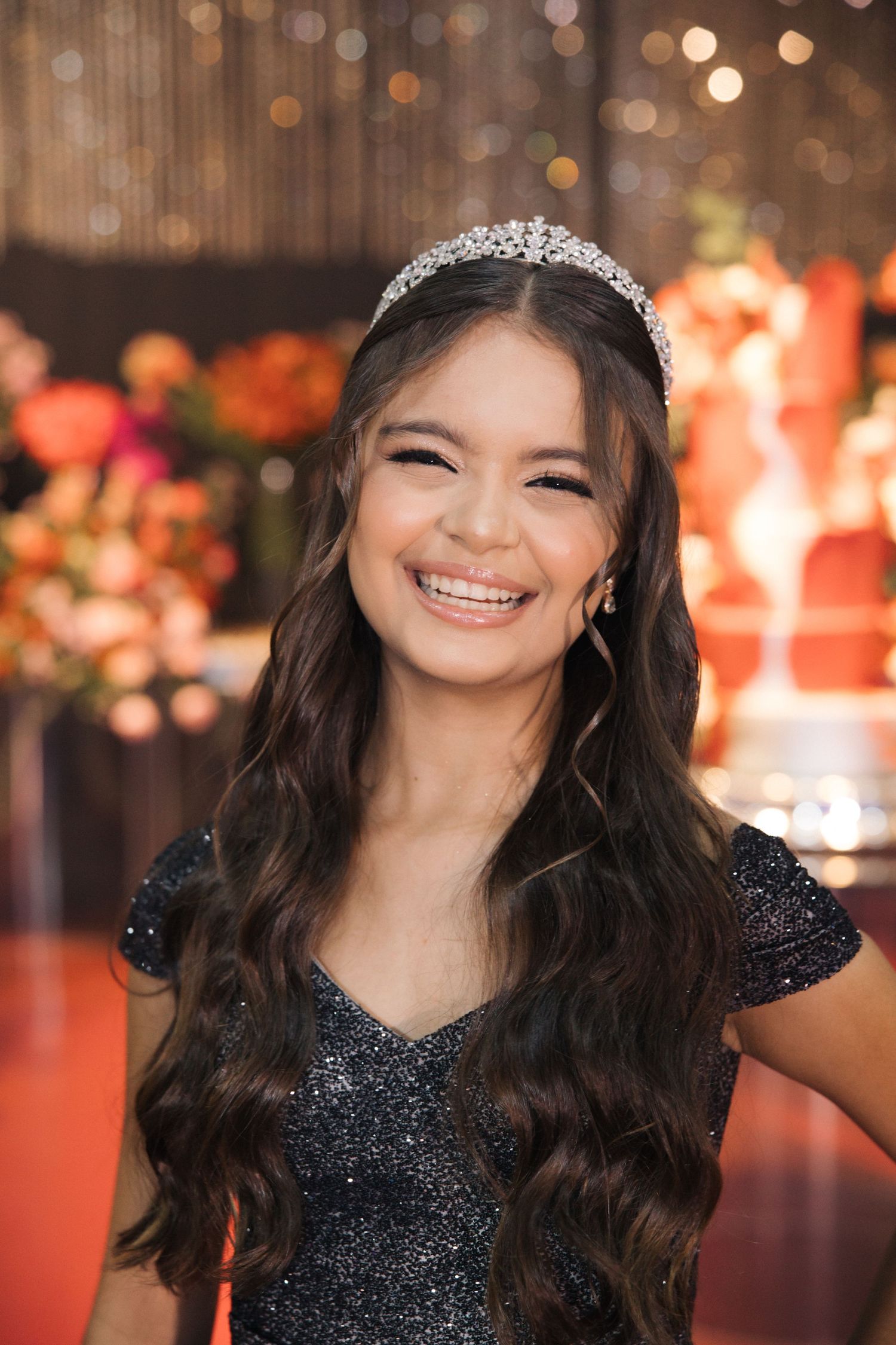 A close-up shot with tiara and sparkly black dress against bokeh lights and floral backdrop.