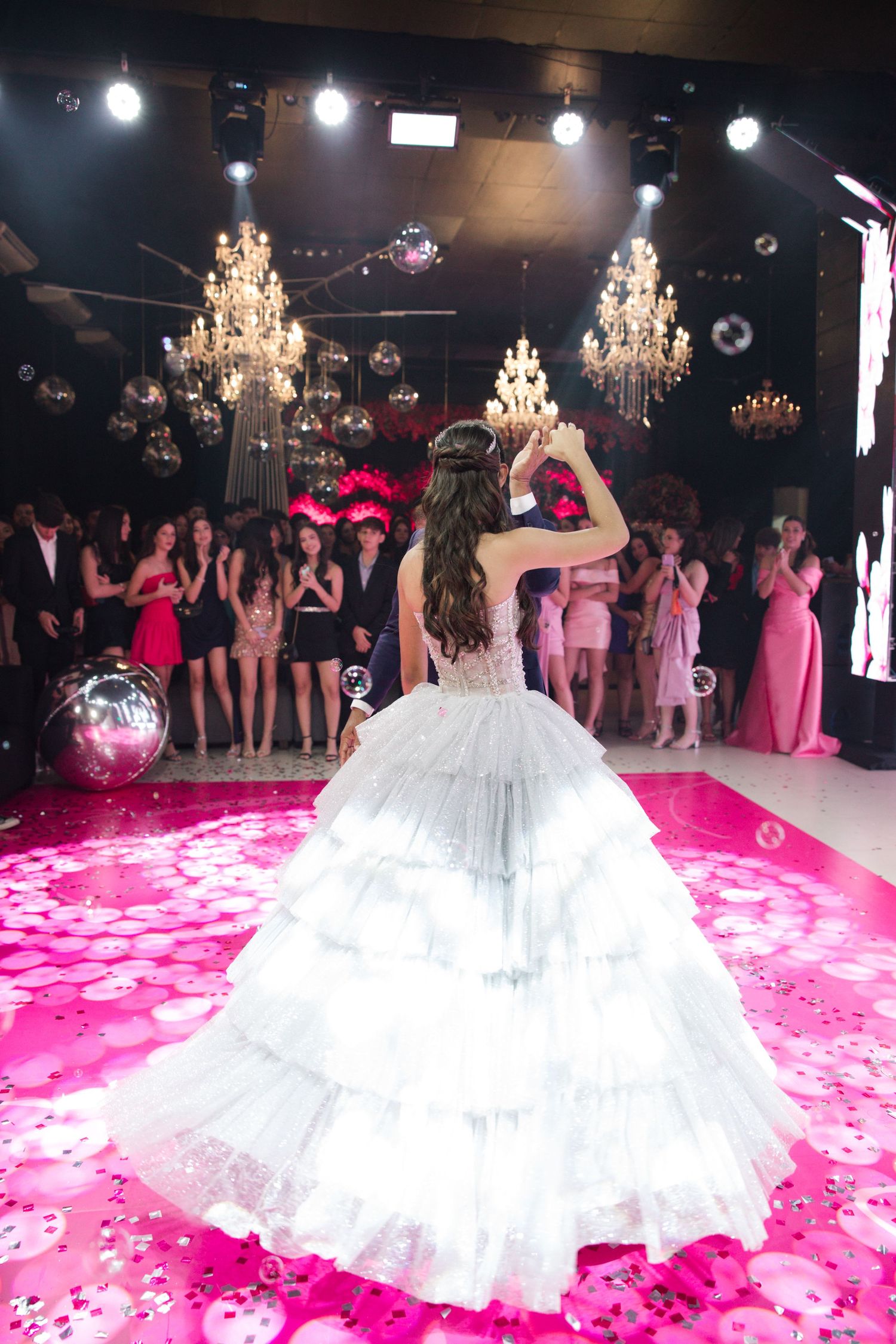 Back view of silver ballgown on pink-lit dance floor with chandeliers and crowd in background.