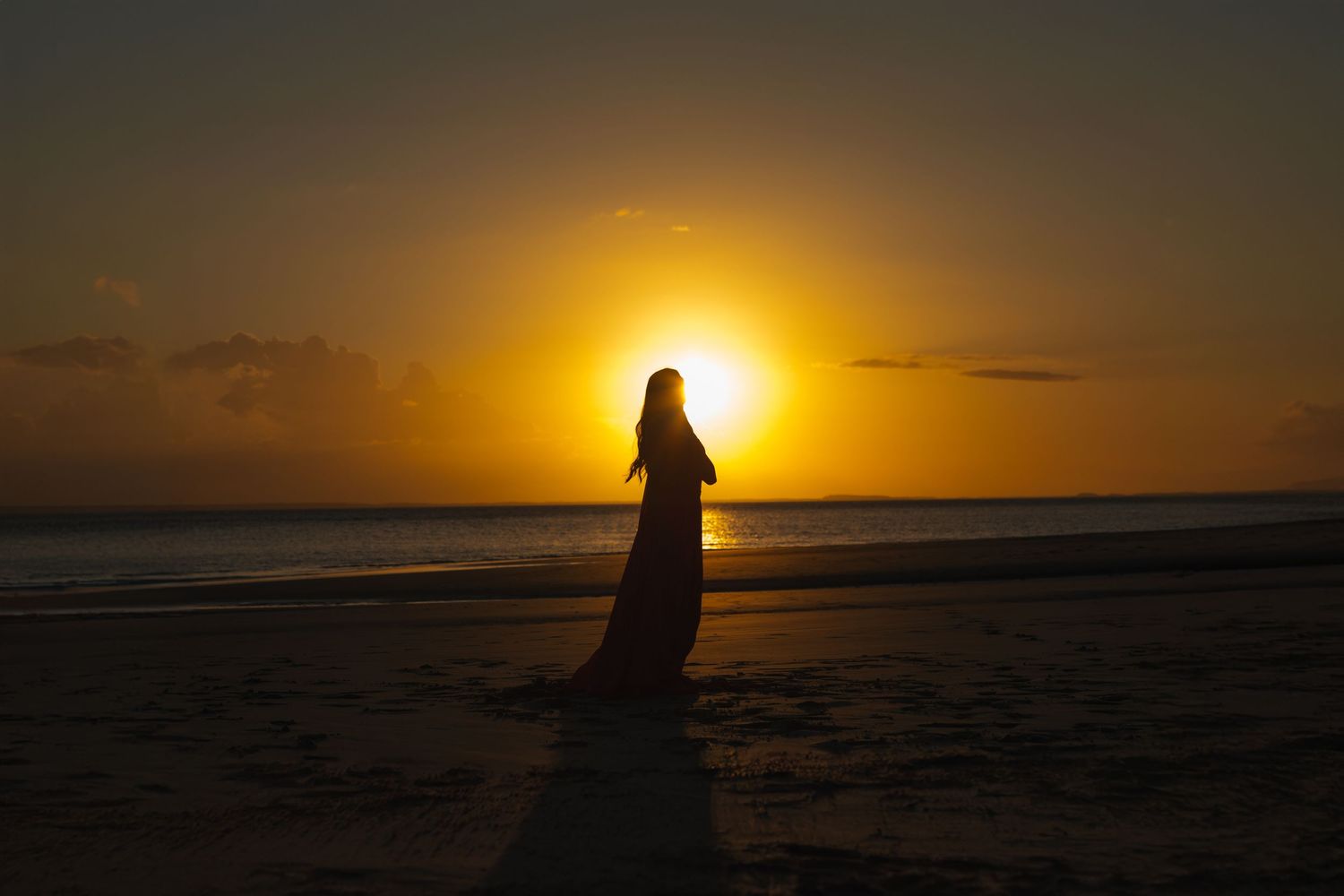 A silhouetted figure in a flowing gown stands on a beach during golden sunset.