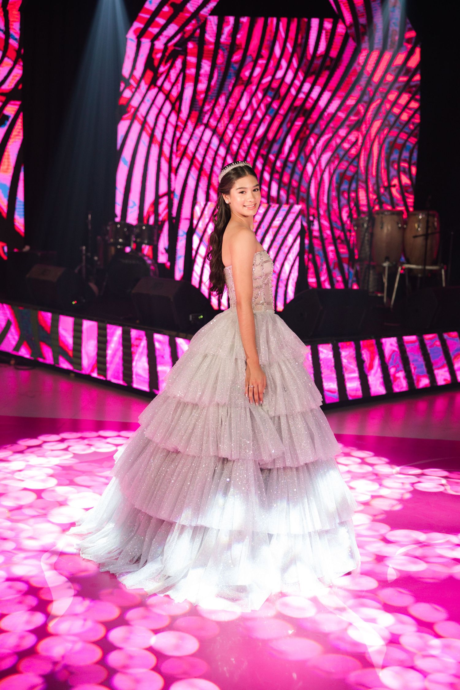 A person wears a flowing layered silver tulle ball gown while posing on a bright pink illuminated stage floor.