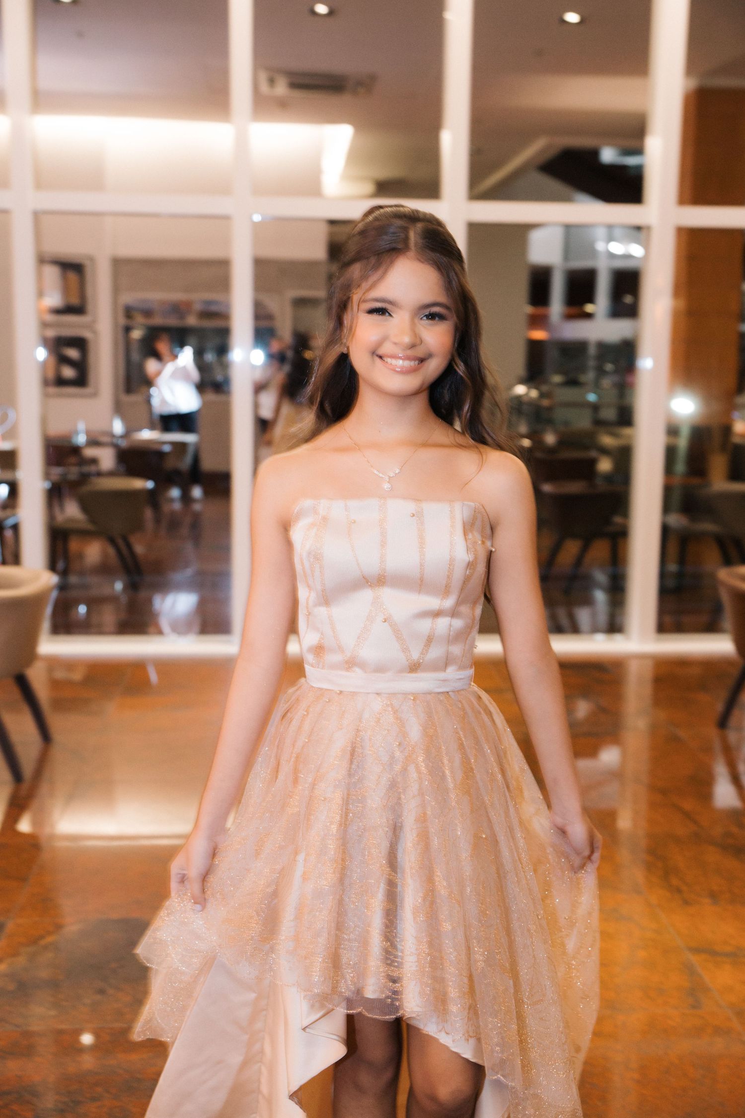 A person in a strapless blush pink tulle dress stands inside a restaurant with hardwood floors and glass windows.
