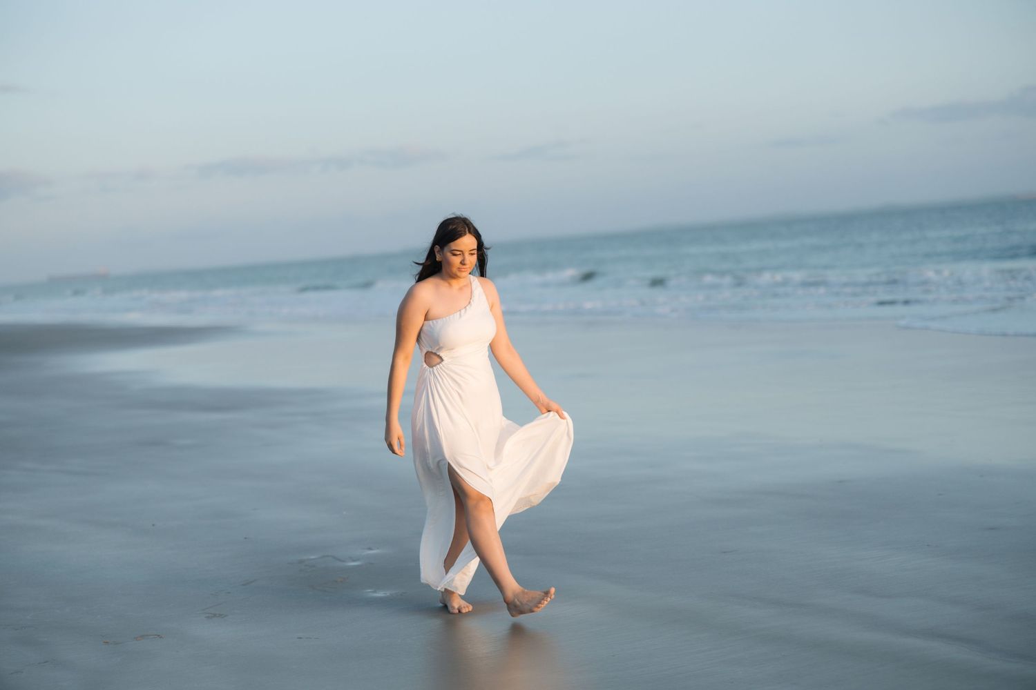 Person in flowing white dress walking barefoot along misty beach shore at dusk.