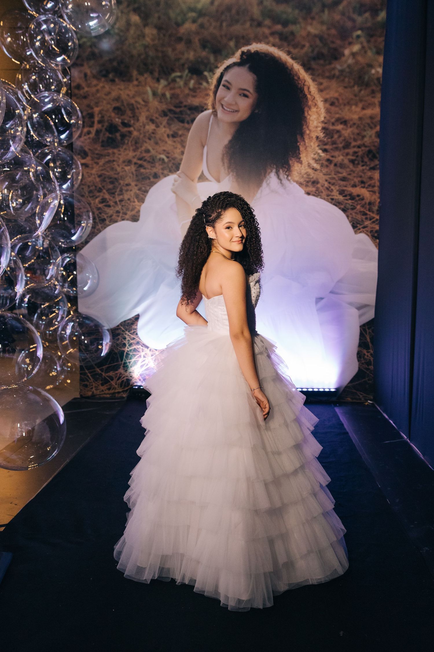 A flowing white tulle ballgown dress with tiered ruffles photographed against a dark backdrop with decorative bubbles.