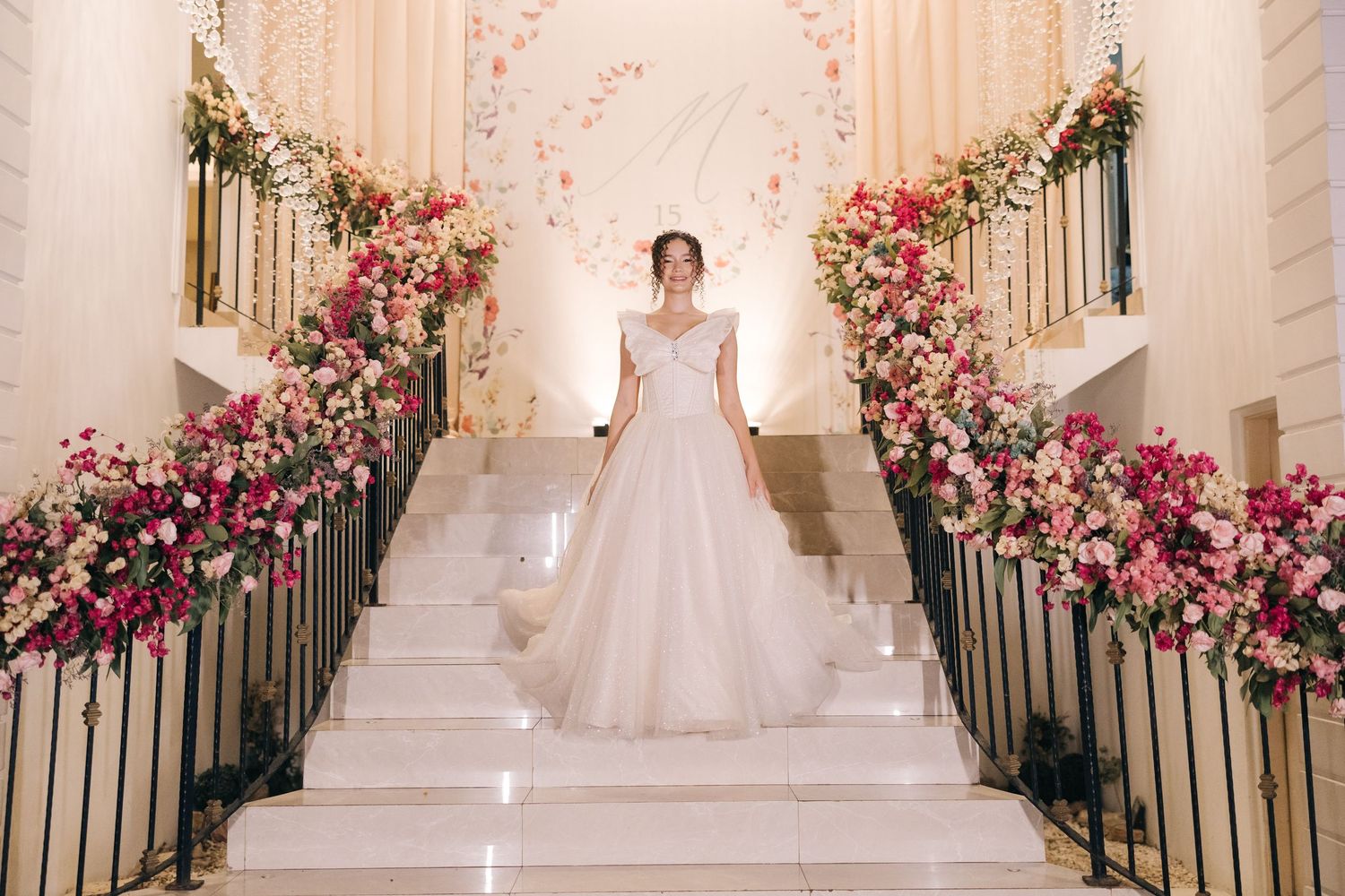 A bride stands elegantly atop a grand staircase adorned with pink and white floral arrangements and gold railings.