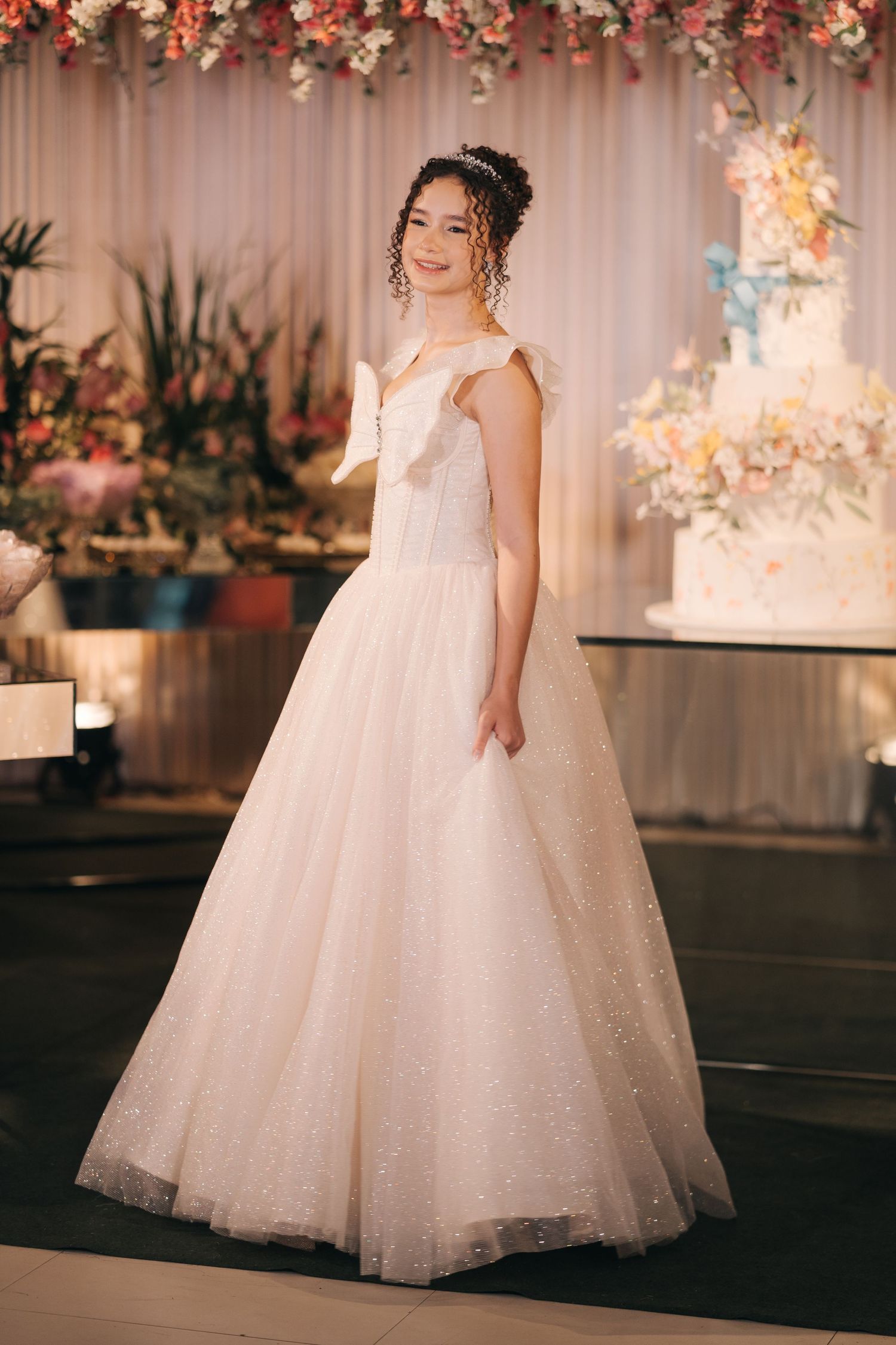 Bride in a stunning white tulle ballgown poses against a floral backdrop at wedding reception.