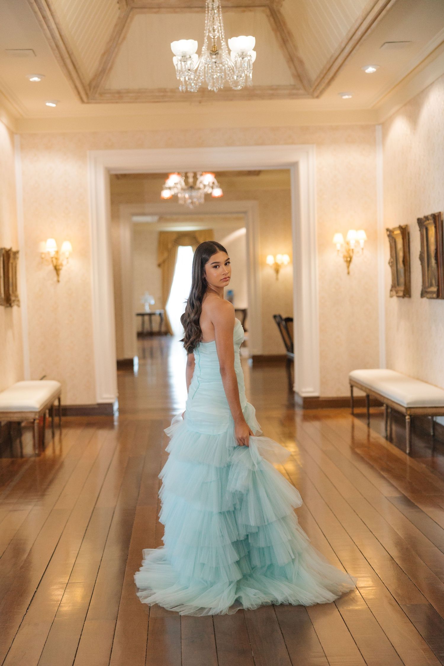 A flowing mint green gown with tiered tulle skirt photographed in an elegant hallway with chandeliers.