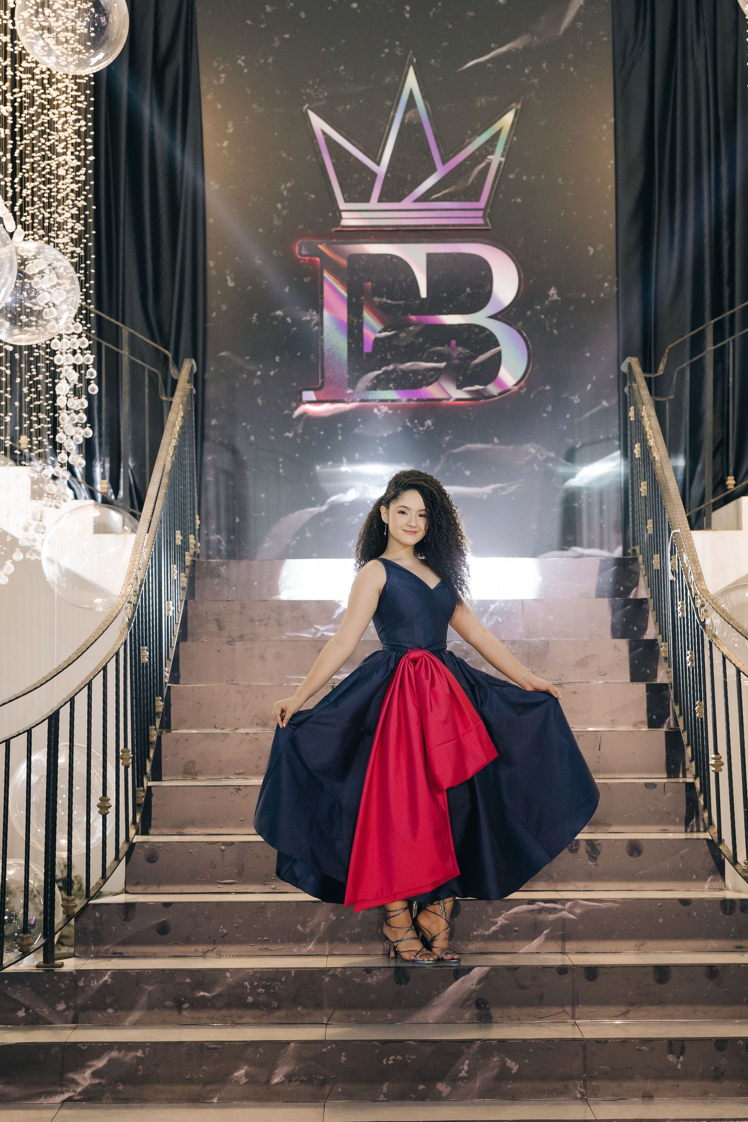 A dramatic navy and red ballgown photographed on grand stairs with a crown logo backdrop and crystal decorations.