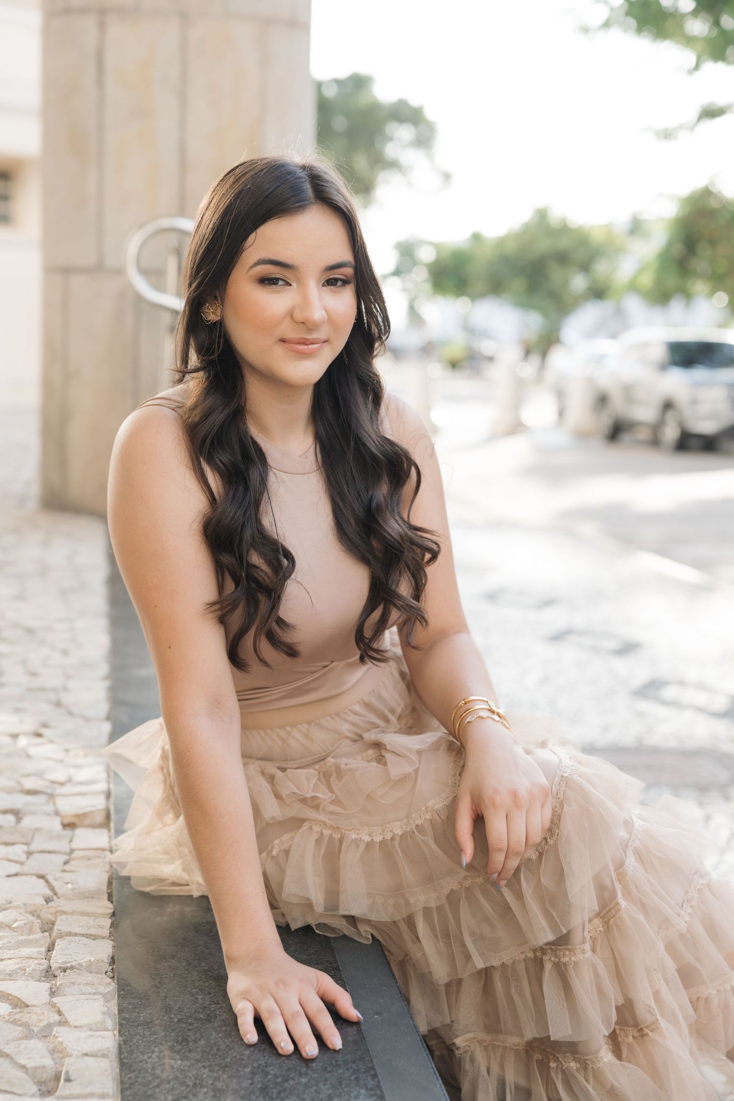 Woman in beige ruffled dress sitting on stone ledge with long dark wavy hair outdoors.