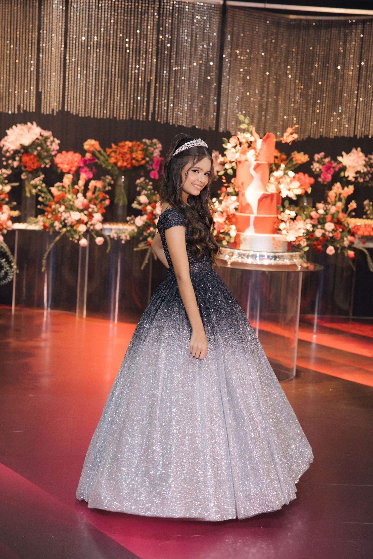 A stunning ombre ball gown transitions from dark to silver sparkles on a red-lit dance floor with floral decorations.