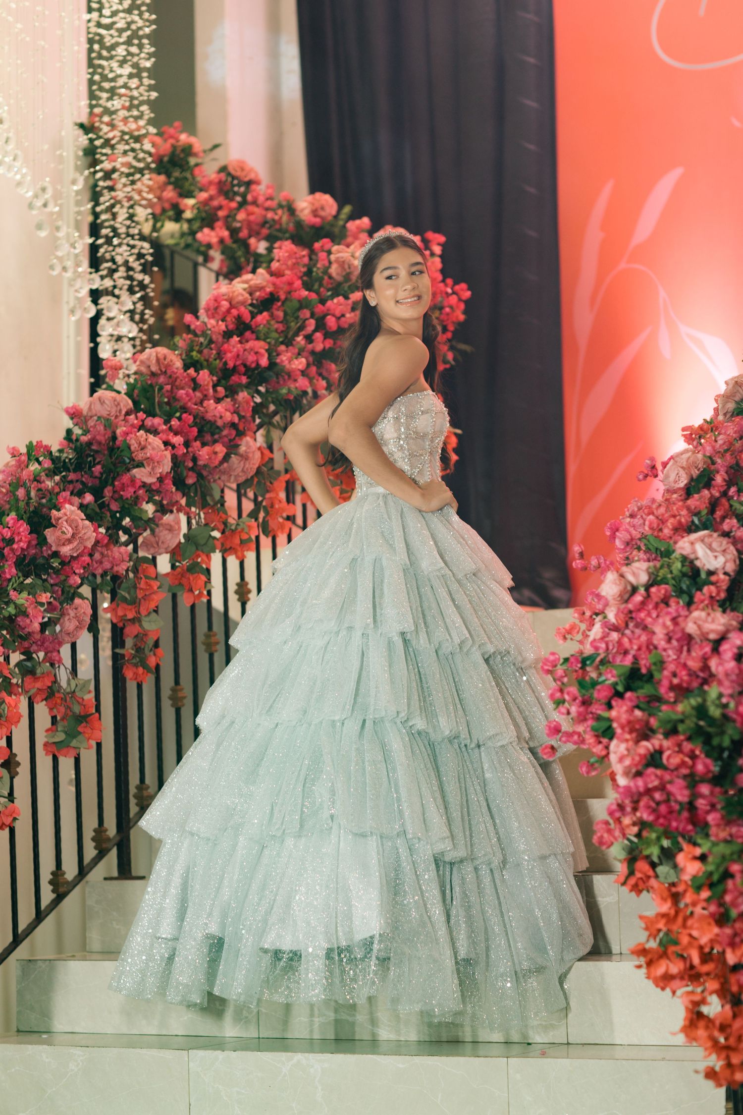 Person in a mint tiered ball gown poses against a backdrop of pink roses and string lights.