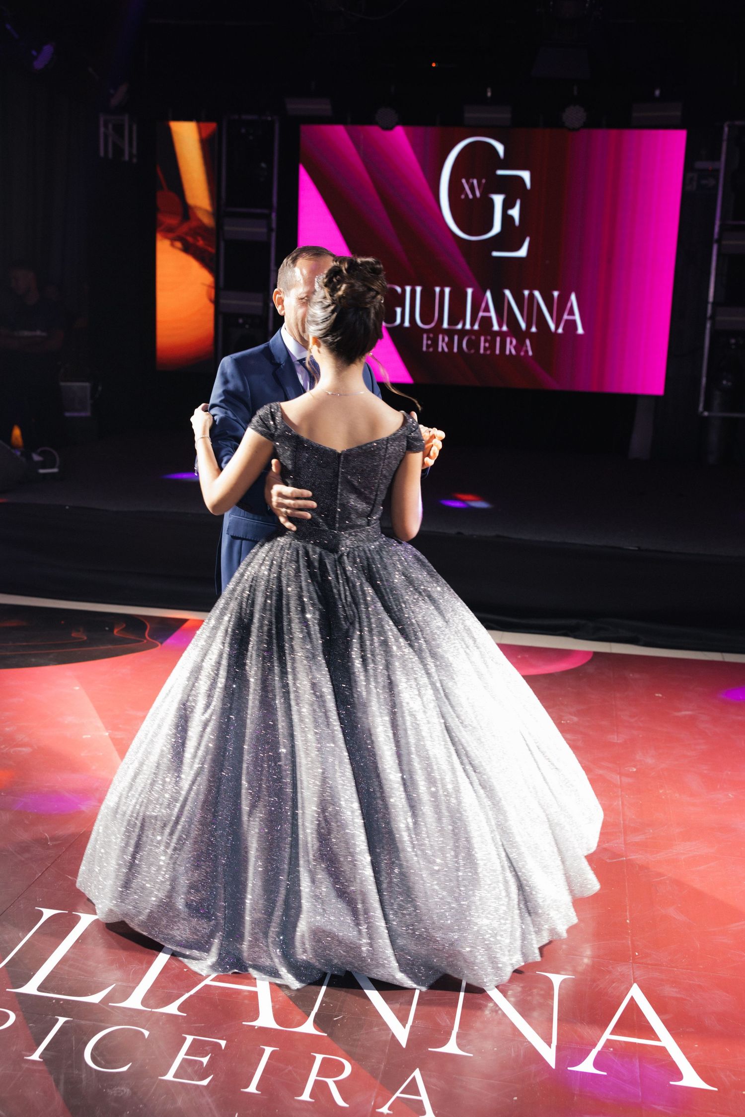 A dancing couple in formal wear seen from behind against a pink-lit backdrop with branded signage.